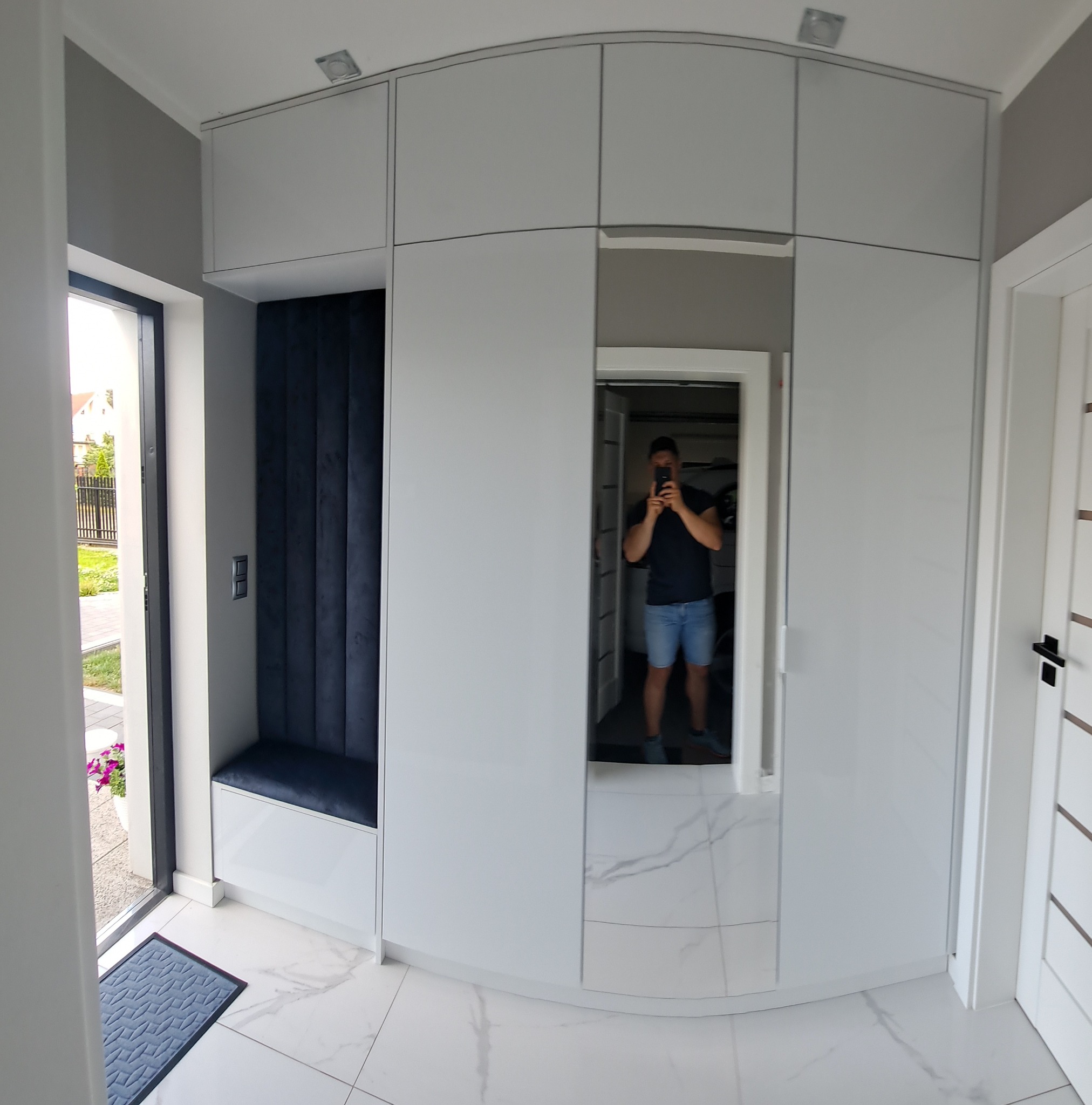 Biała szafa wnękowa z siedziskiem i lustrem, odbijającym postać. Nowoczesny design, jasne wnętrze, marmurowe płytki podłogowe. Widok z perspektywy wejścia.