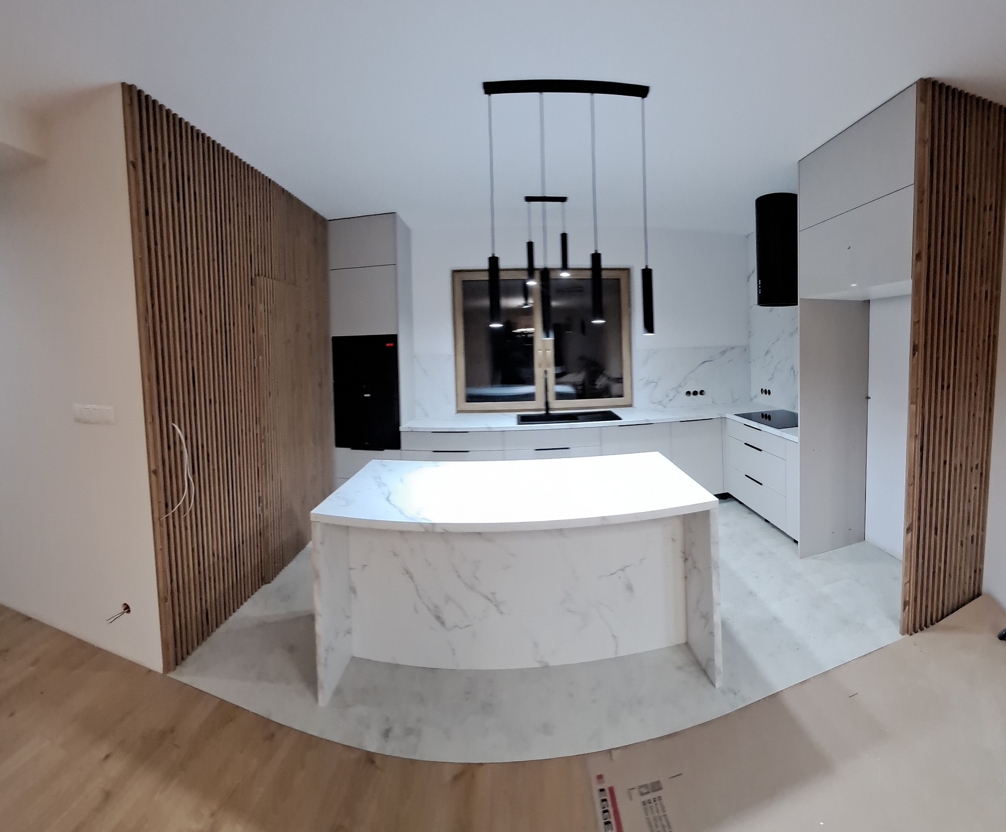 Nowoczesna kuchnia z wyspą i drewnianymi lamelami na ścianie. Białe szafki, czarny okap i lampy nad wyspą. Marmurowy wzór na wyspie i ścianie.