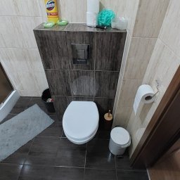 SPAWTECH KRÓL SPÓŁKA Z OGRANICZONĄ ODPOWIEDZIALNOŚCIĄ - Nowoczesna toaleta z zabudowaną spłuczką i pojemnikiem na środki czystości. Widoczny fragment kabiny prysznicowej i dywanik łazienkowy. Ujęcie z góry.