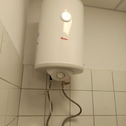 SPAWTECH KRÓL SPÓŁKA Z OGRANICZONĄ ODPOWIEDZIALNOŚCIĄ - Biały, wiszący bojler elektryczny marki Mister, zamontowany na kafelkowanej ścianie w łazience. Widoczne przyłącza wodne i kabel zasilający.