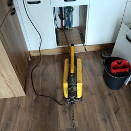 SPAWTECH KRÓL SPÓŁKA Z OGRANICZONĄ ODPOWIEDZIALNOŚCIĄ - Elektryczna przepychaczka do rur pod zlewozmywakiem z otwartymi drzwiczkami szafki, widoczne rury. Obok stoi czarne wiadro z czerwoną wkładką.