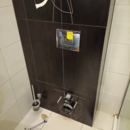 SPAWTECH KRÓL SPÓŁKA Z OGRANICZONĄ ODPOWIEDZIALNOŚCIĄ - Podtynkowa instalacja WC w łazience z brązowymi płytkami. Widoczne elementy montażowe, narzędzia i fragment odbicia osoby w chromowanym panelu.