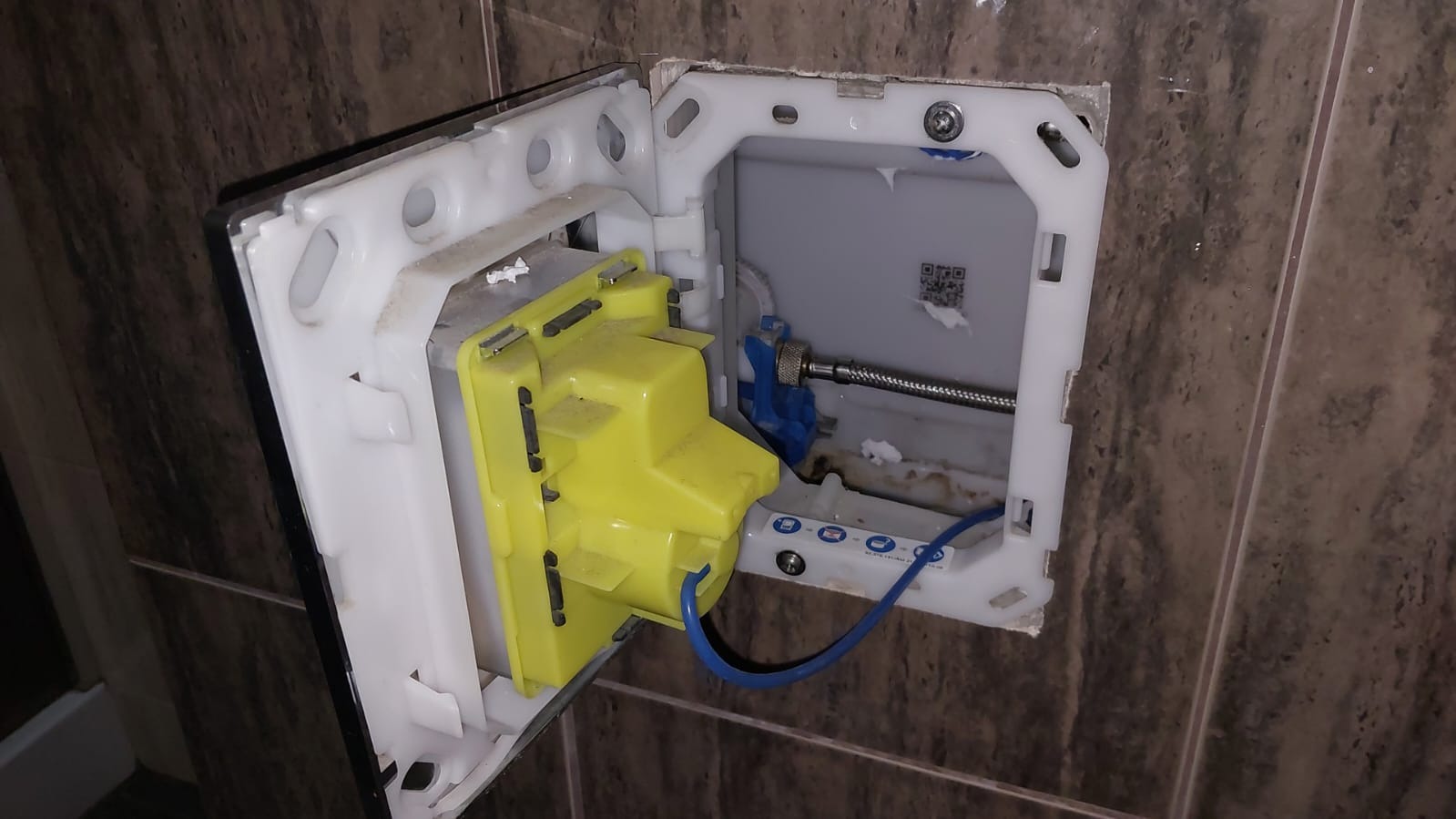 Otwarta obudowa podtynkowa WC z widocznym mechanizmem spłukującym w kolorze żółtym i niebieskim, zamontowana na brązowej ścianie wyłożonej płytkami.