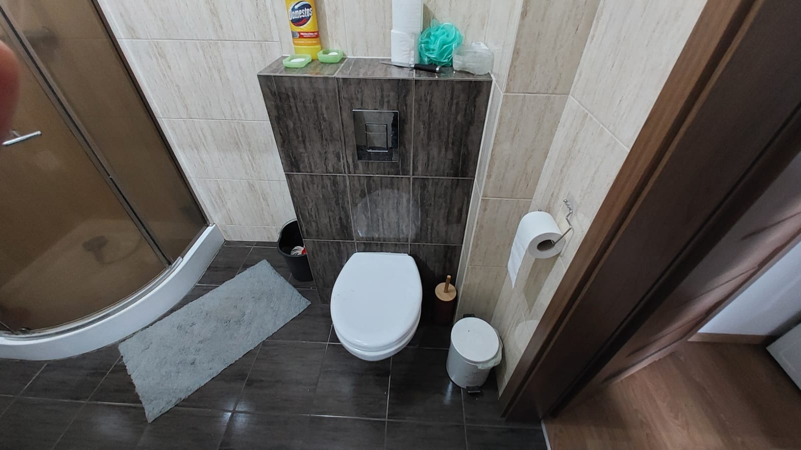 Nowoczesna toaleta z zabudowaną spłuczką i pojemnikiem na środki czystości. Widoczny fragment kabiny prysznicowej i dywanik łazienkowy. Ujęcie z góry.