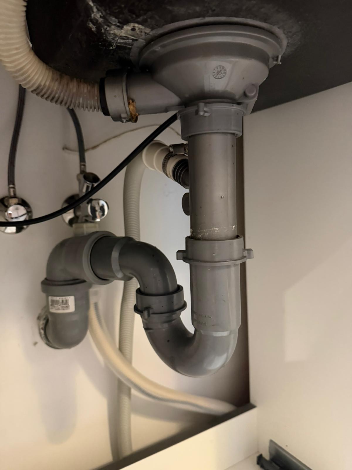 Szare rury odpływowe pod zlewem, widoczne złącza i wężyki. Instalacja hydrauliczna w szafce, z elementami mocującymi i uszczelniającymi. Detal hydrauliczny.