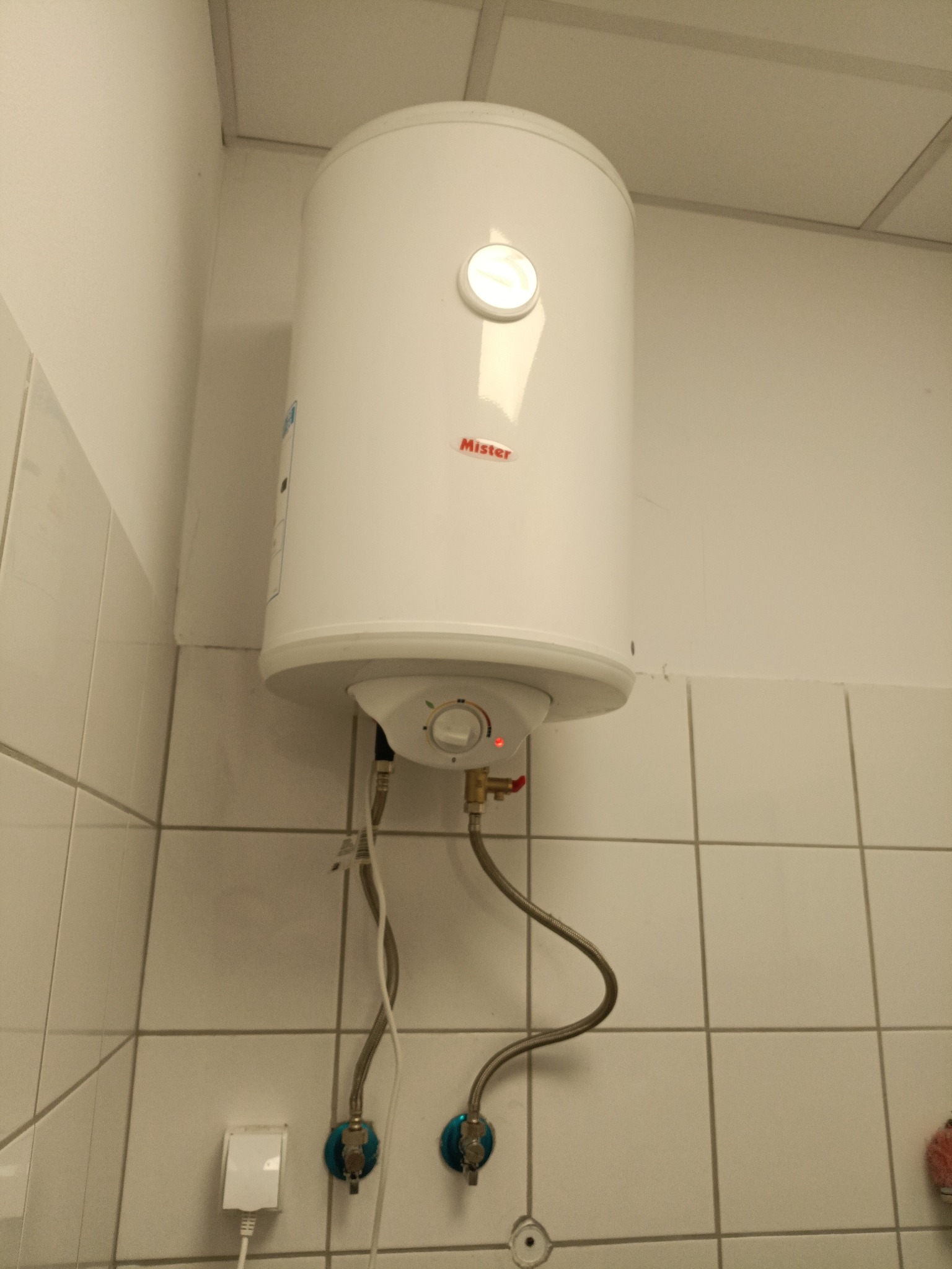 Biały, wiszący bojler elektryczny marki Mister, zamontowany na kafelkowanej ścianie w łazience. Widoczne przyłącza wodne i kabel zasilający.