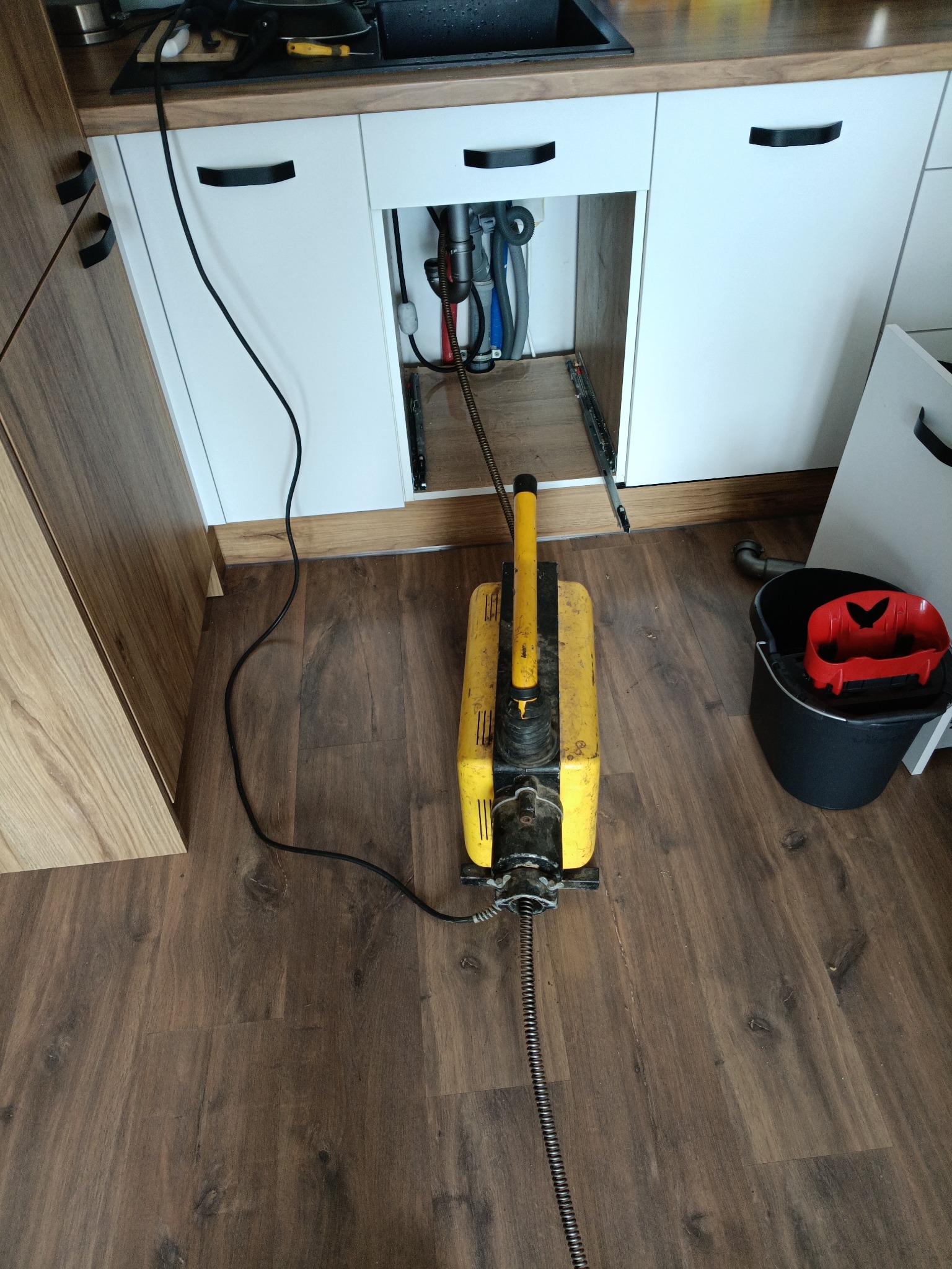 Elektryczna przepychaczka do rur pod zlewozmywakiem z otwartymi drzwiczkami szafki, widoczne rury. Obok stoi czarne wiadro z czerwoną wkładką.