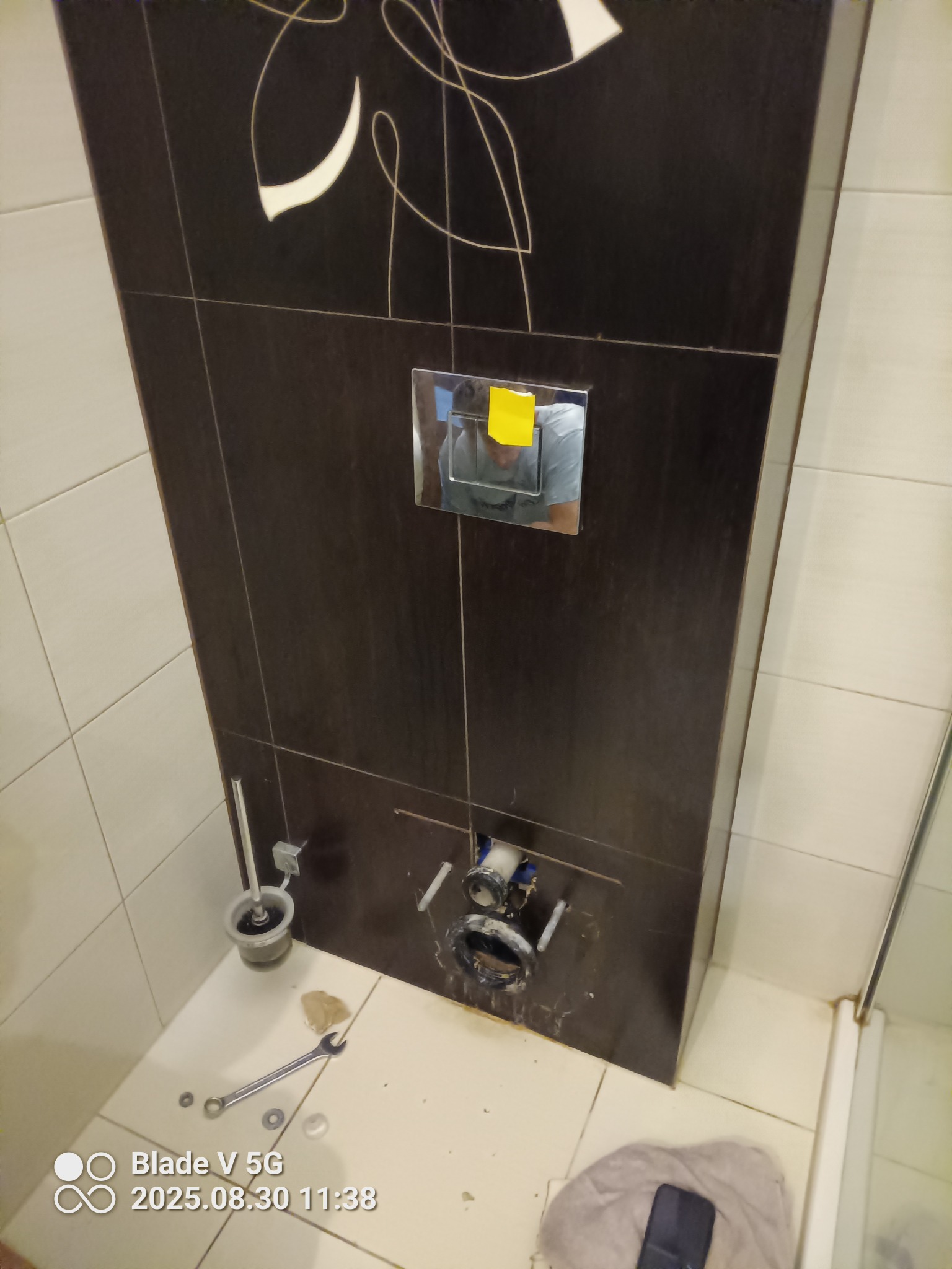 Podtynkowa instalacja WC w łazience z brązowymi płytkami. Widoczne elementy montażowe, narzędzia i fragment odbicia osoby w chromowanym panelu.