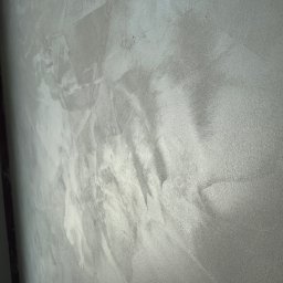 RIWIA SPÓŁKA Z OGRANICZONĄ ODPOWIEDZIALNOŚCIĄ - Szara ściana z efektem dekoracyjnym, imitującym beton. Widoczne nieregularne wzory i faktura. Ujęcie z boku, podkreślające strukturę powierzchni.