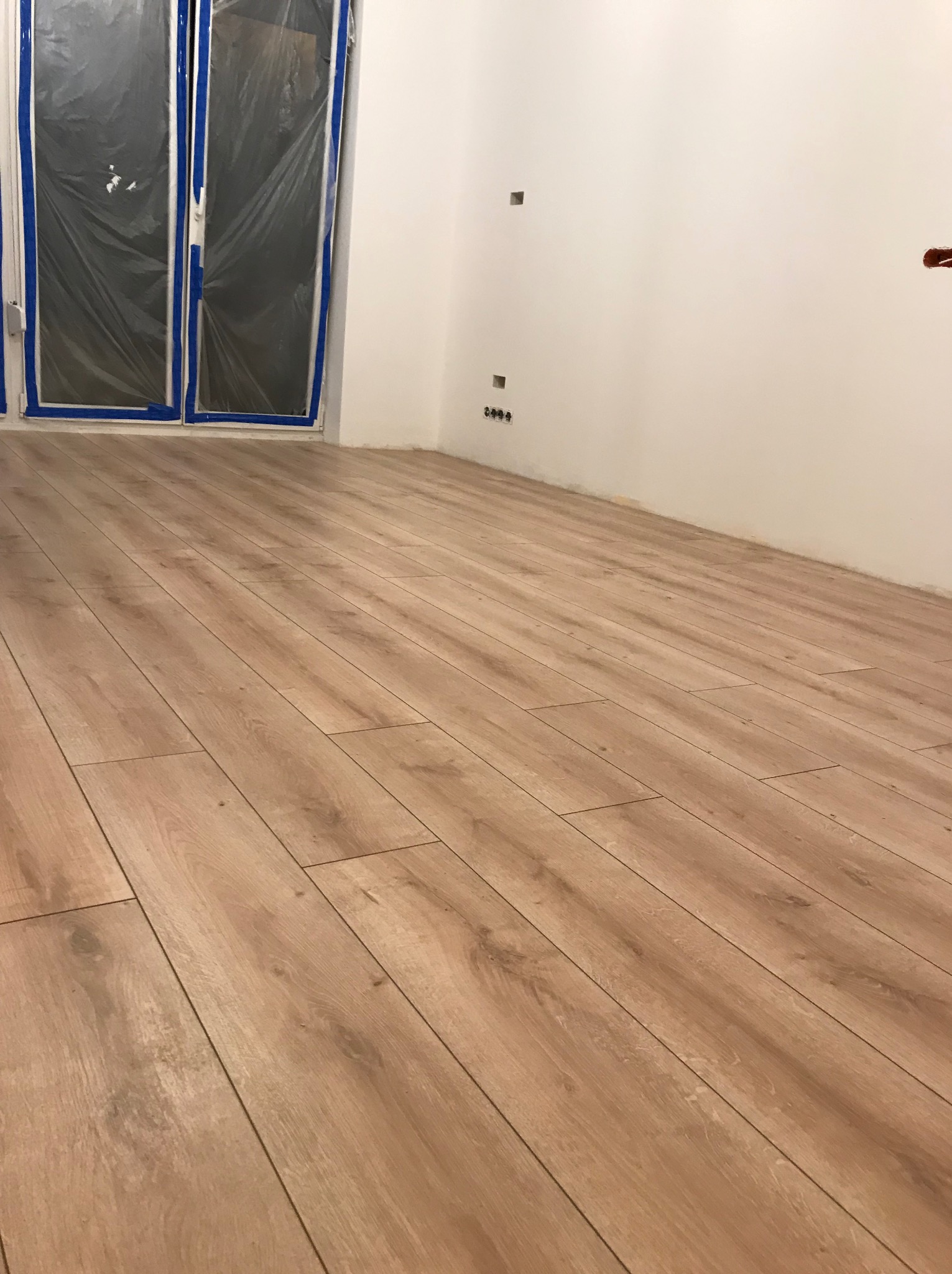 Jasne panele podłogowe ułożone w pokoju z białymi ścianami i zabezpieczonymi folią oknami. Widoczne gniazdka elektryczne. Ujęcie z niskiej perspektywy.