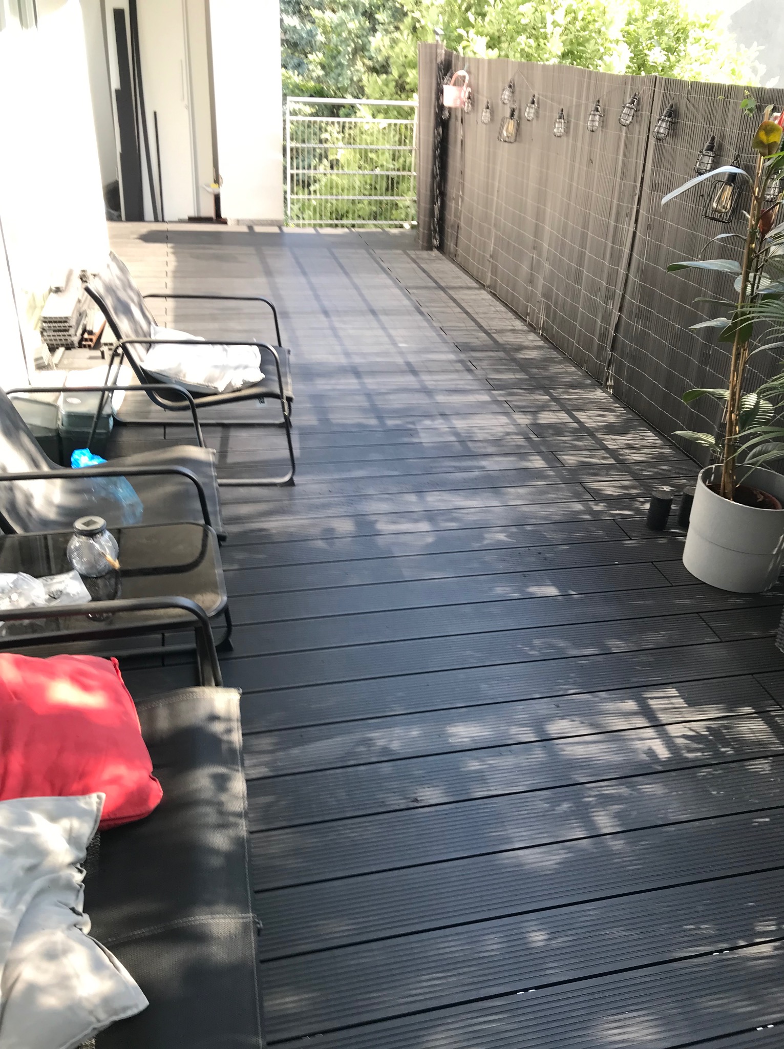 Nowoczesny balkon z szarymi panelami podłogowymi, metalowymi krzesłami i dekoracyjnym oświetleniem. Widok na zieleń za balustradą. Cień drzew na podłodze.