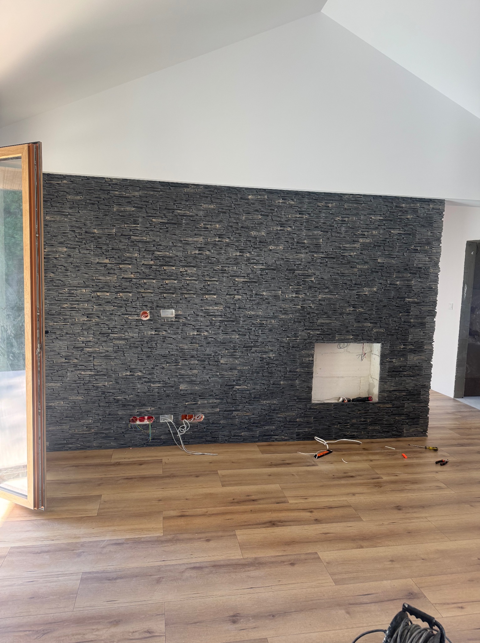 Wykończone wnętrze z panelami podłogowymi i ścianą z kamienia dekoracyjnego. Widoczne gniazdka elektryczne i narzędzia, co sugeruje etap prac wykończeniowych.