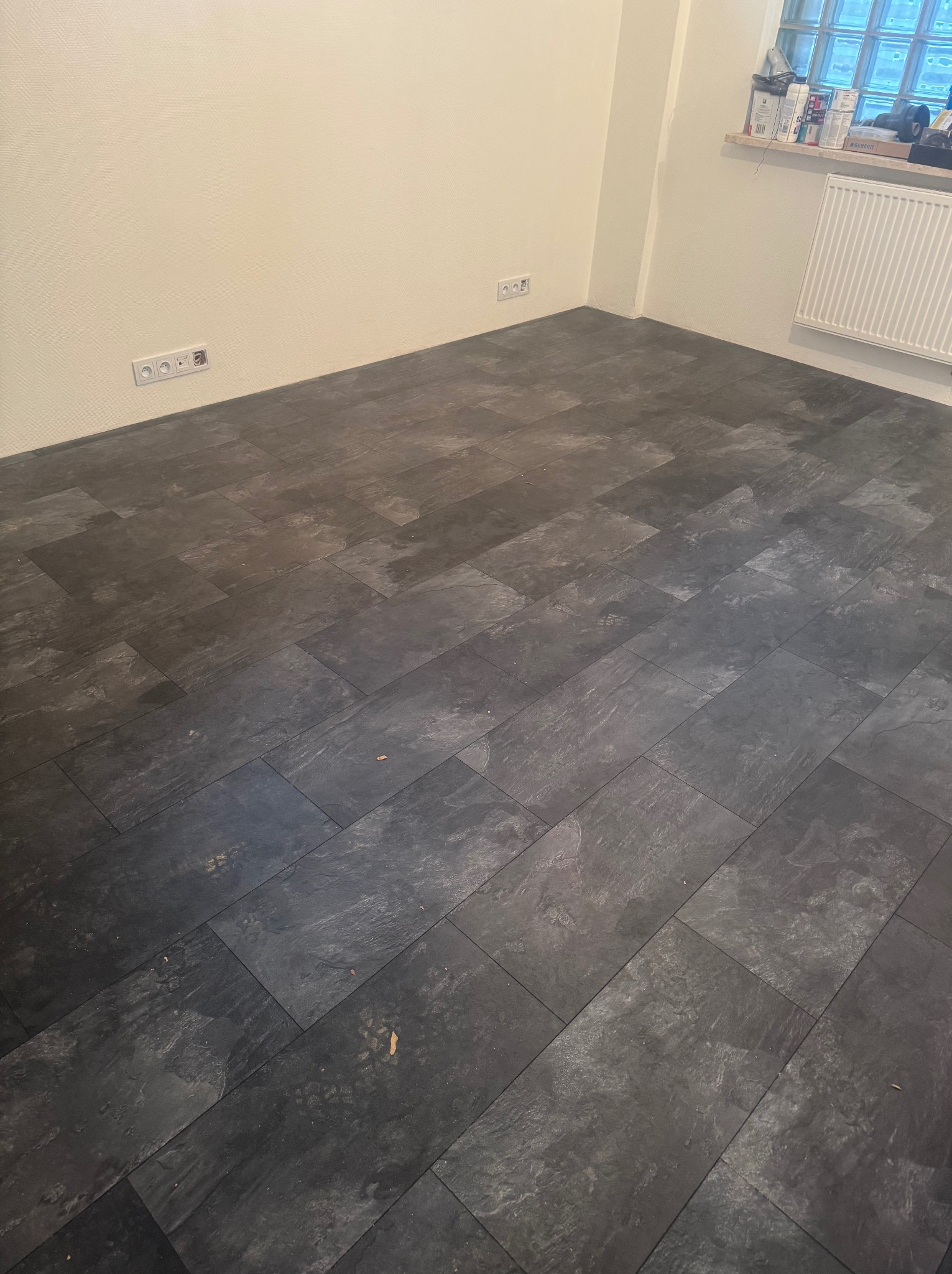 Ukończona podłoga z ciemnych paneli imitujących kamień w rogu pokoju z białymi ścianami i grzejnikiem. Widoczne gniazdka elektryczne i parapet z przedmiotami.