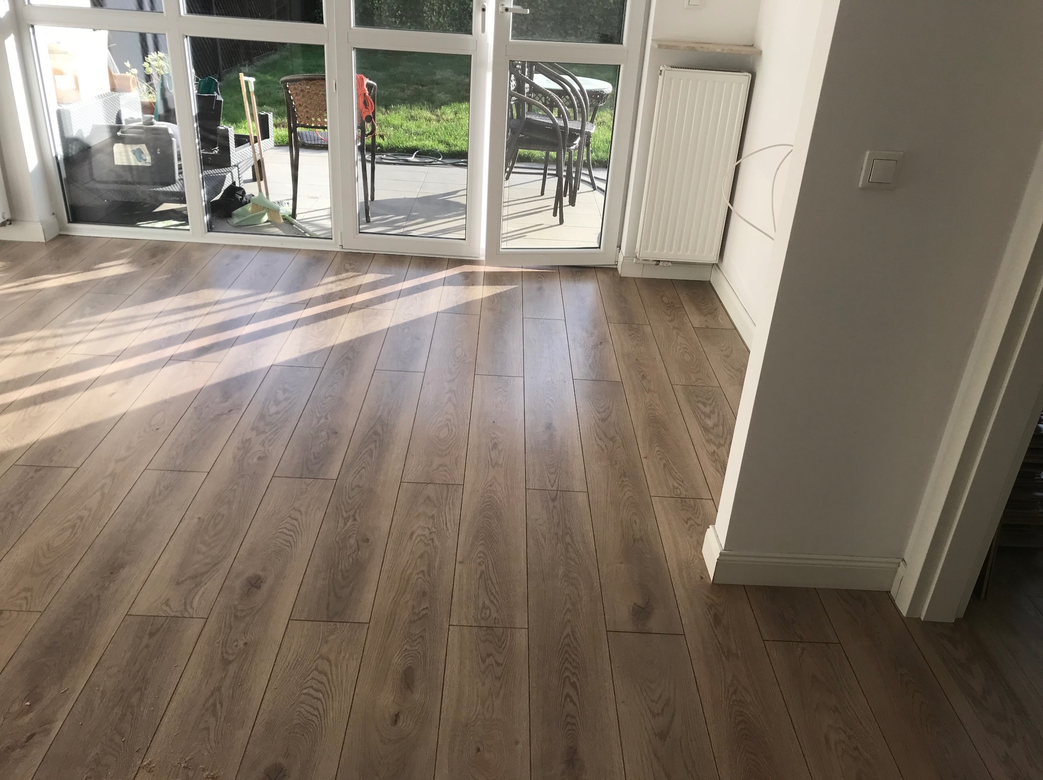 Jasne panele podłogowe ułożone wzdłuż okna balkonowego, widoczne detale tekstury drewna i subtelne refleksy światła. Białe listwy przypodłogowe i fragment grzejnika.