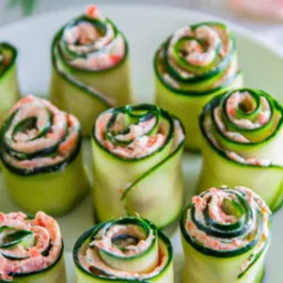 Dekoracyjne roladki z cukinii wypełnione kremowym farszem na białym talerzu, idealne jako przekąska na przyjęciach. Widoczne gałązki koperku w tle.