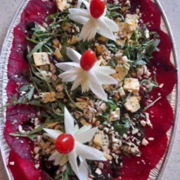 Owalna taca z folii aluminiowej prezentuje kompozycję z plastrów buraka, rukoli, sera pleśniowego, orzechów i balsamicznego dressingu, ozdobioną kwiatami z warzyw i pomidorkami.