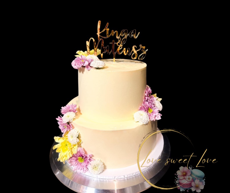 Dwupoziomowy tort w kremowym kolorze, udekorowany świeżymi kwiatami i złotym topperem z imionami, na czarnym tle. Idealny na uroczystość.