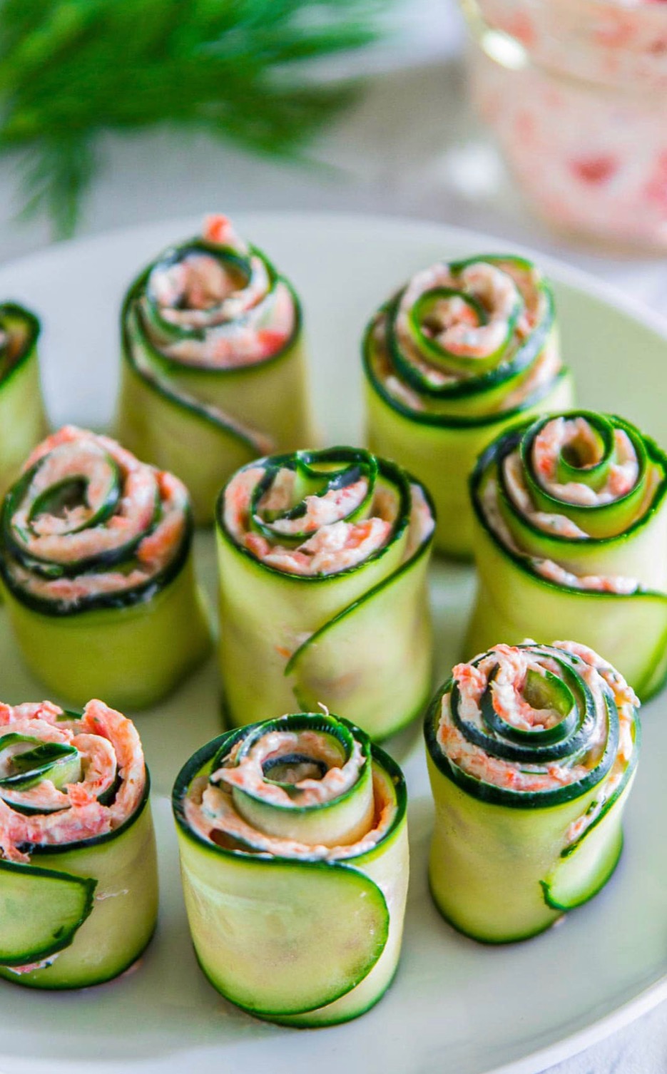 Dekoracyjne roladki z cukinii wypełnione kremowym farszem na białym talerzu, idealne jako przekąska na przyjęciach. Widoczne gałązki koperku w tle.