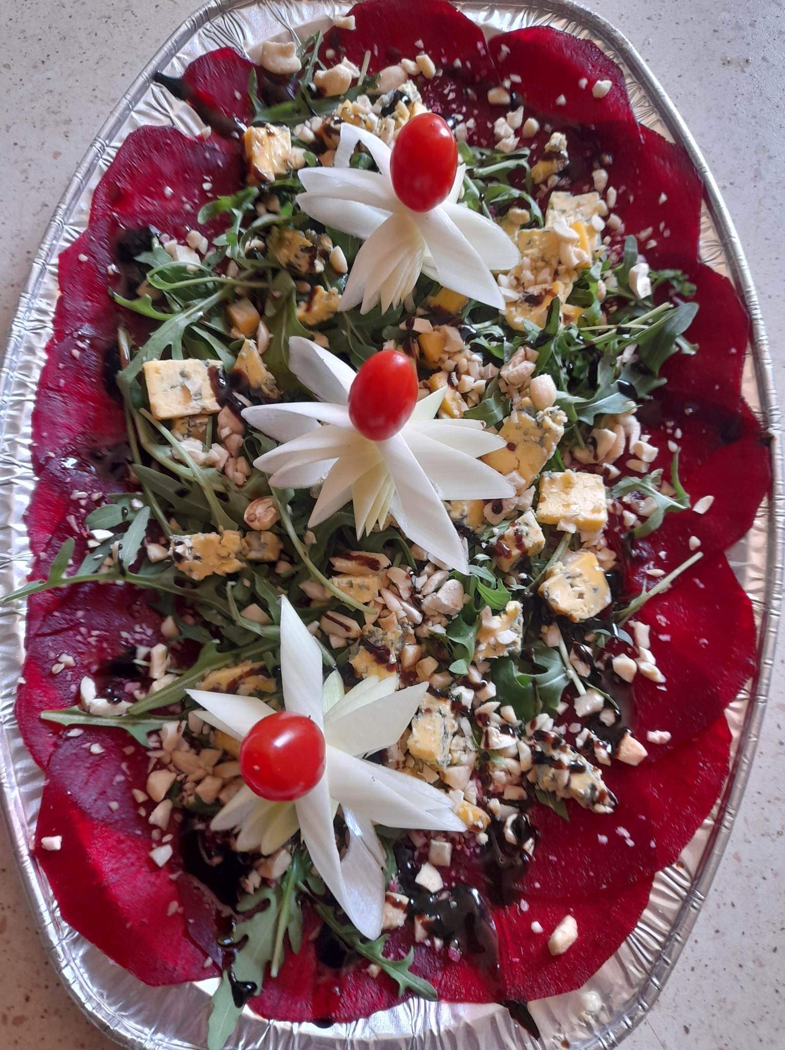Owalna taca z folii aluminiowej prezentuje kompozycję z plastrów buraka, rukoli, sera pleśniowego, orzechów i balsamicznego dressingu, ozdobioną kwiatami z warzyw i pomidorkami.