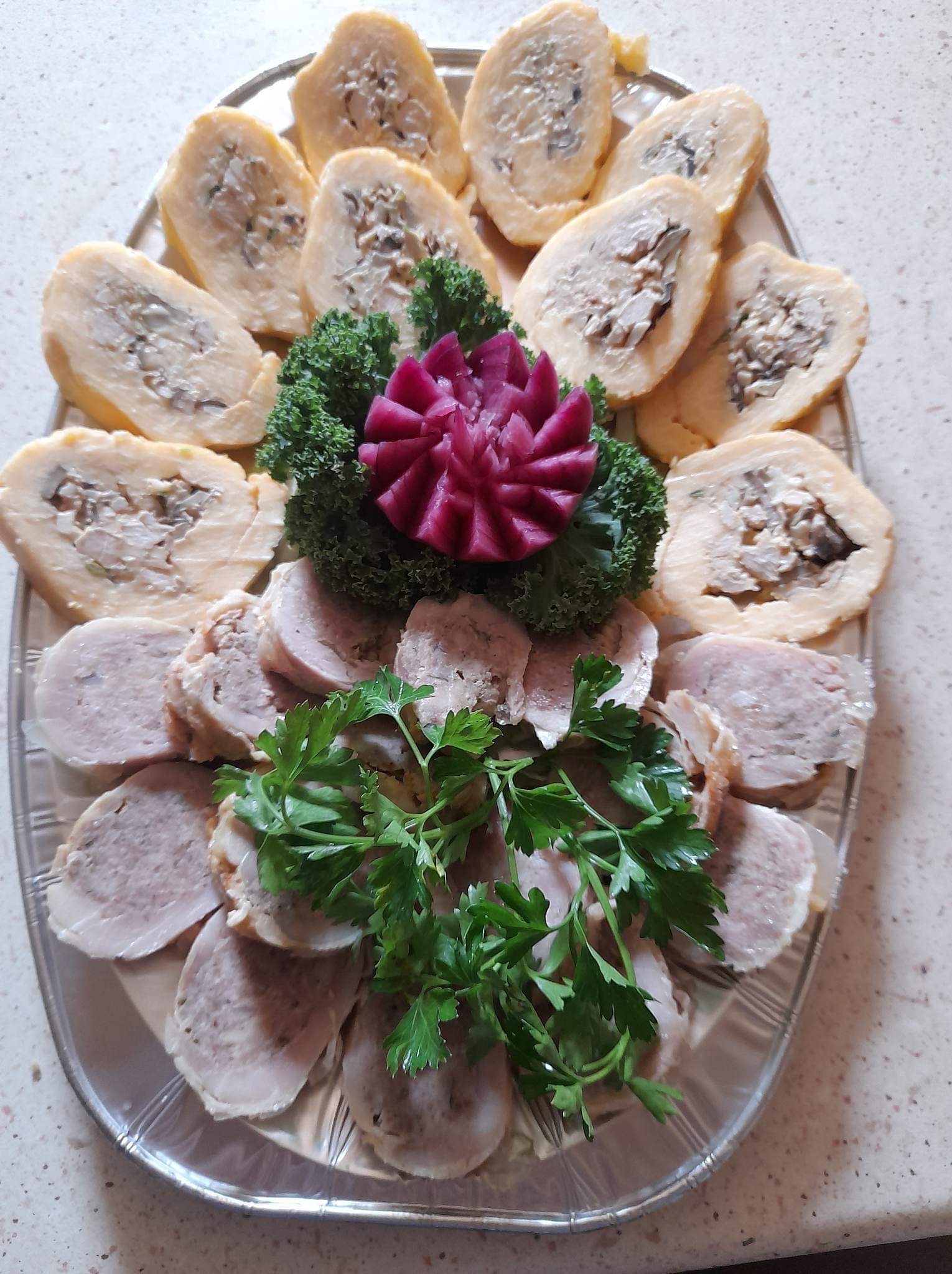 Dekoracyjna taca z roladkami mięsnymi i jajecznymi, ozdobiona zieloną pietruszką i rzeźbionym burakiem, prezentowana na jasnym tle.