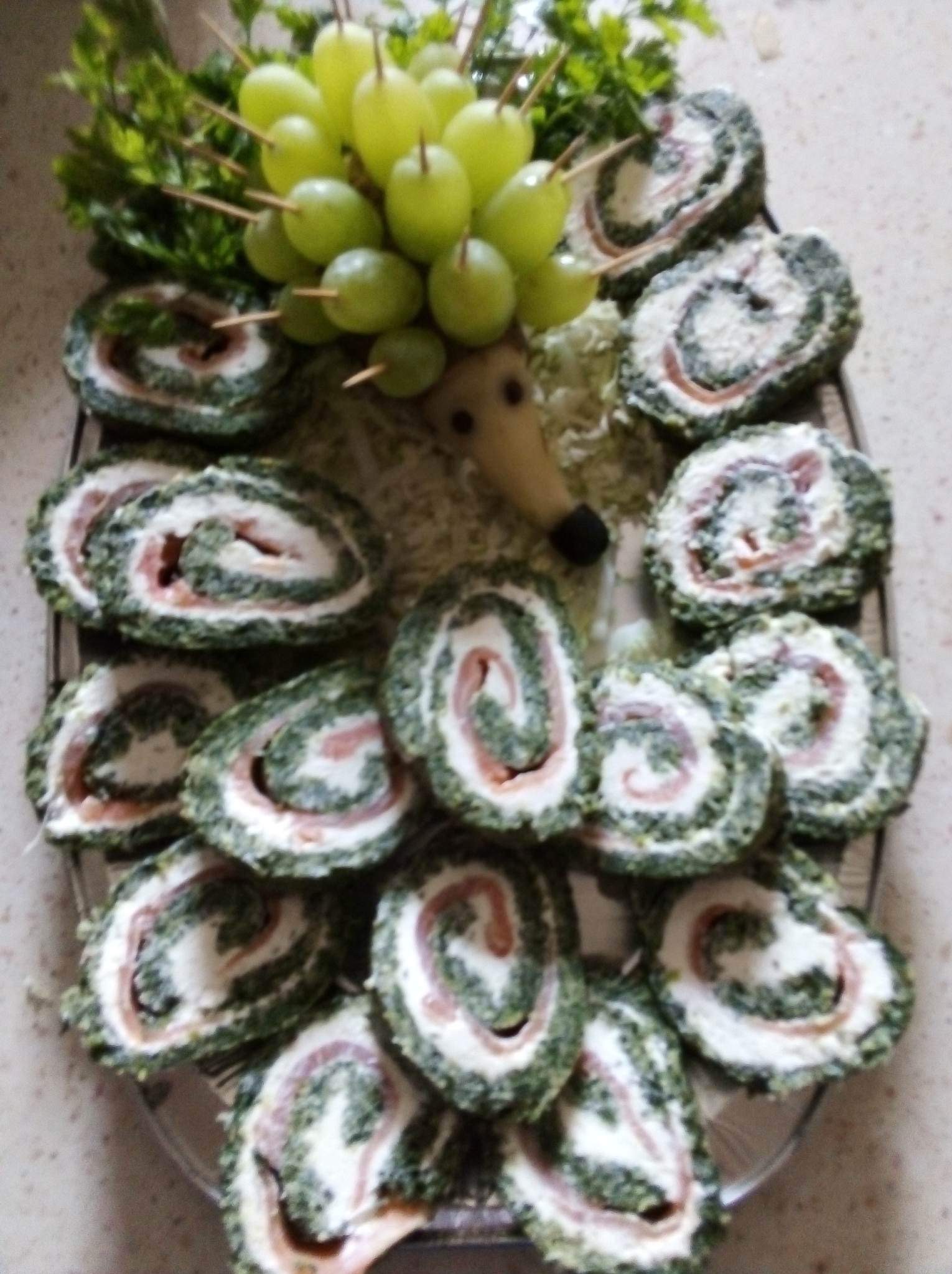 Dekoracyjna rolada szpinakowa z łososiem na paterze, ułożona w kształt jeża z winogronami i natką pietruszki. Kreatywne danie na imprezę.
