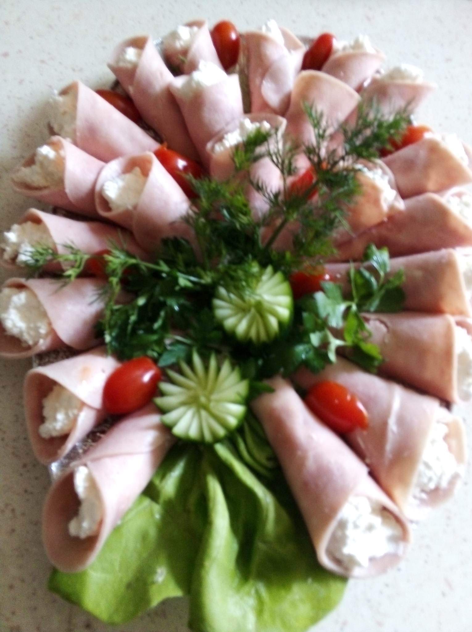 Elegancko ułożone roladki z szynki z kremowym nadzieniem, udekorowane pomidorkami cherry, świeżym koperkiem i rzeźbionym ogórkiem na liściu sałaty.
