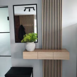 Nowoczesna konsola z lustrem i lamelami ściennymi w przedpokoju. Jasne drewno, czarne akcenty, minimalistyczny design. Roślina w doniczce dodaje naturalnego akcentu.