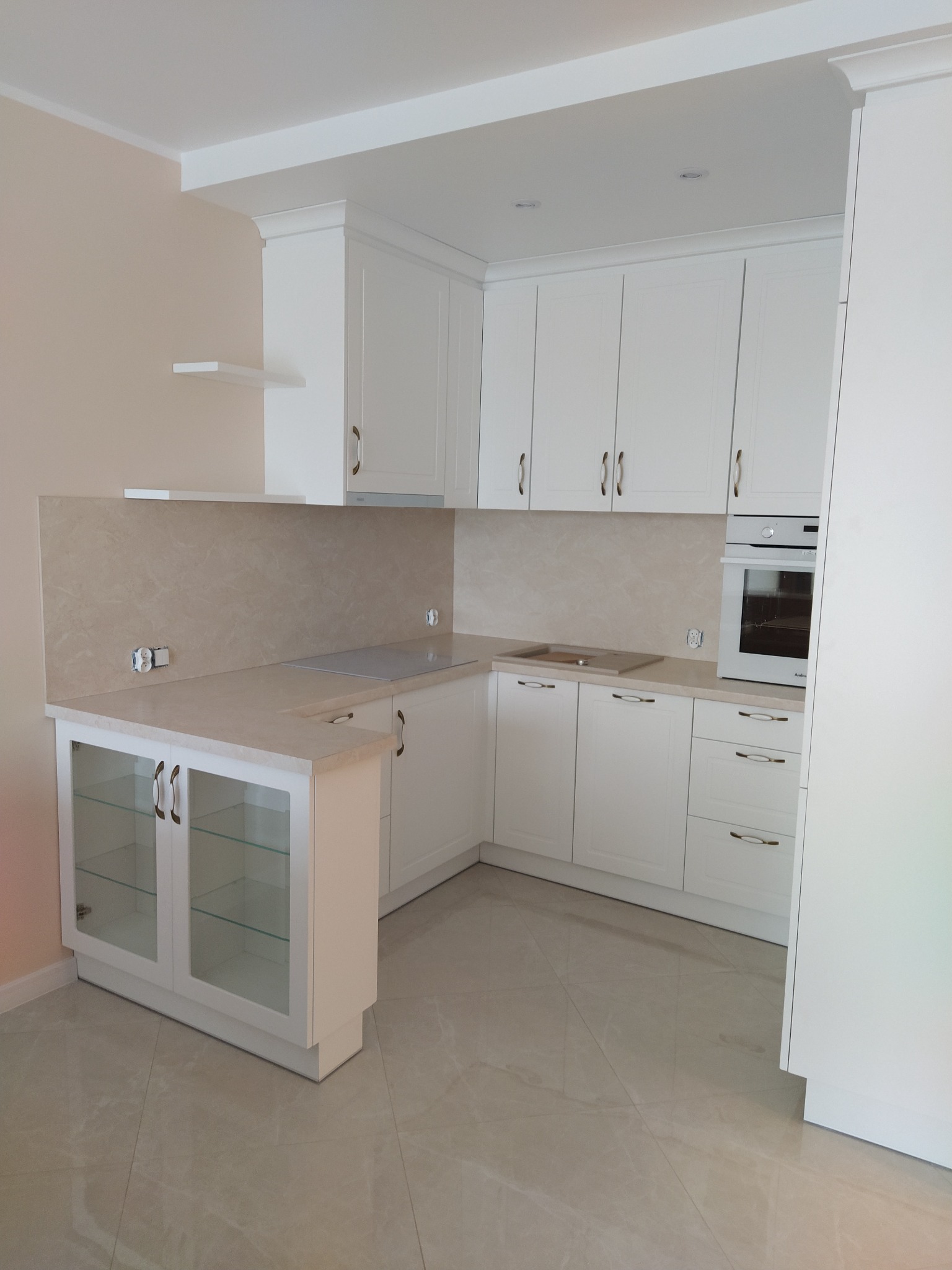 Jasna kuchnia z białymi szafkami, szklanymi frontami i beżowym blatem. Widoczny fragment płyty indukcyjnej i piekarnika. Uwagę zwraca minimalistyczna półka ścienna.