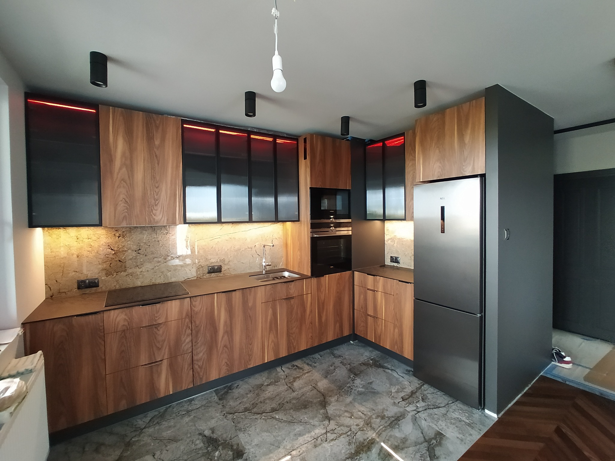 Nowoczesna kuchnia z drewnianymi frontami, czarnymi akcentami i marmurowym blatem. Czerwone podświetlenie górnych szafek. Widoczna lodówka i elementy zabudowy AGD.