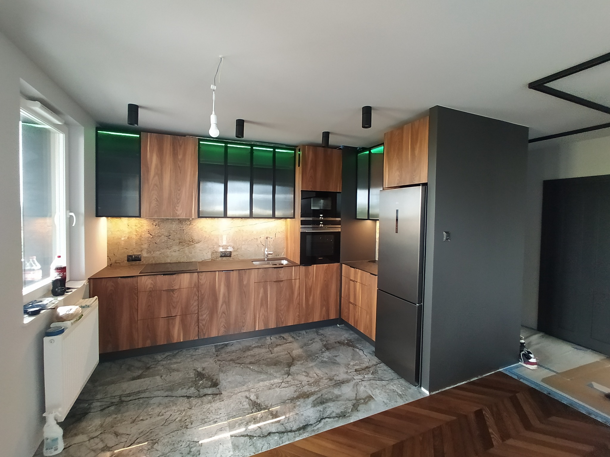 Nowoczesna kuchnia z drewnianymi frontami, czarnymi akcentami i zielonym oświetleniem LED nad szafkami. Marmurowa podłoga i minimalistyczny design.