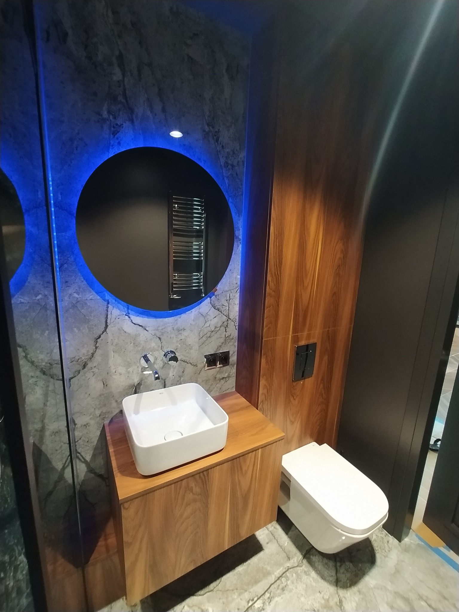 Nowoczesna łazienka z umywalką nablatową na drewnianej szafce, okrągłym lustrem z niebieskim podświetleniem LED i wiszącą toaletą w minimalistycznym stylu.