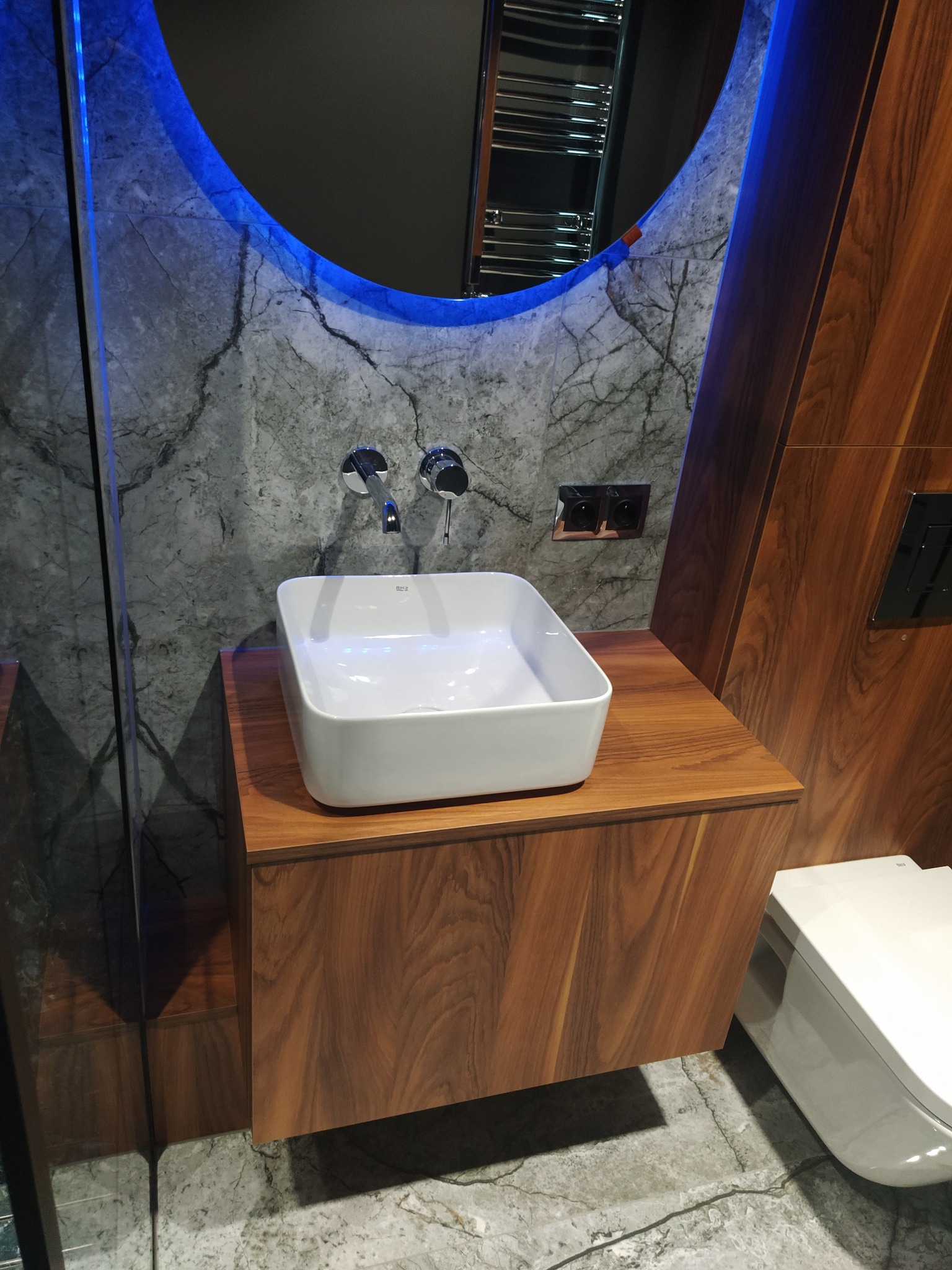 Nowoczesna łazienka: biała, kwadratowa umywalka Roca na szafce z orzecha włoskiego, marmurowe ściany, okrągłe lustro z niebieskim podświetleniem LED.