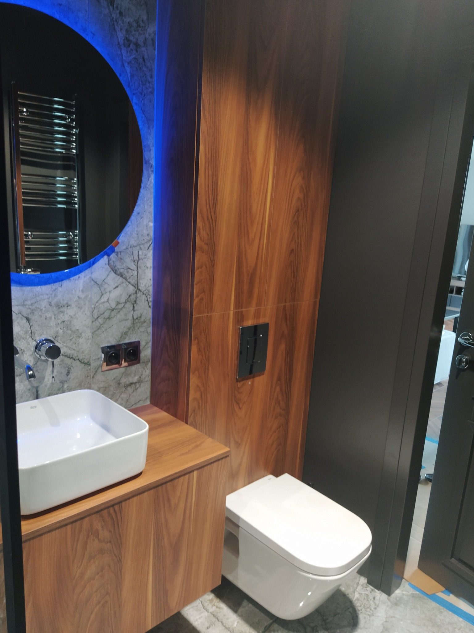 Nowoczesna łazienka z drewnianą szafką pod umywalką i toaletą w zabudowie. Okrągłe lustro z niebieskim podświetleniem i marmurowa ściana dodają elegancji.