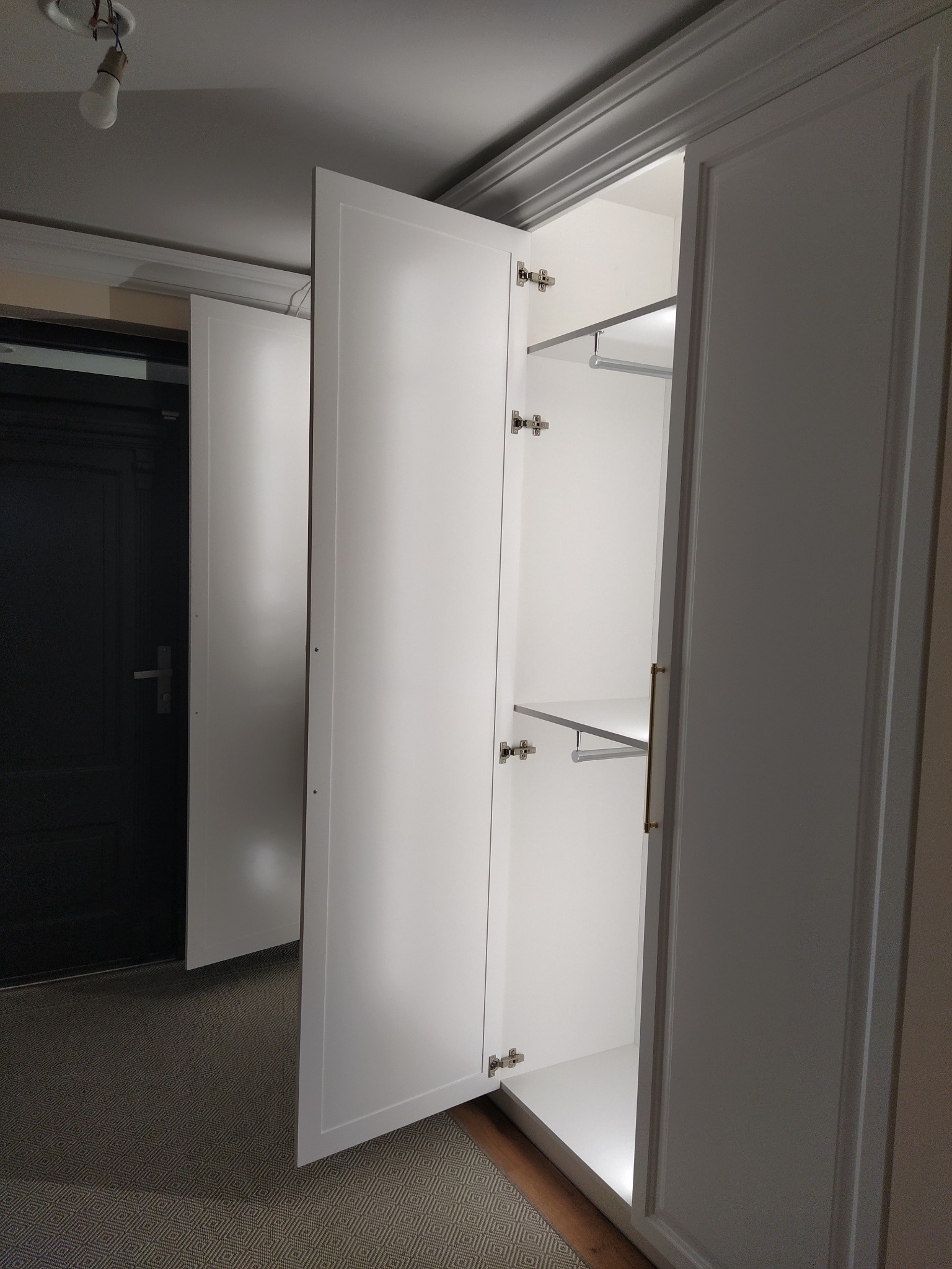 Biała szafa wnękowa z otwartymi drzwiami, widoczne półki i oświetlenie LED, na tle ciemnych drzwi i wzorzystej podłogi. Minimalistyczny design.