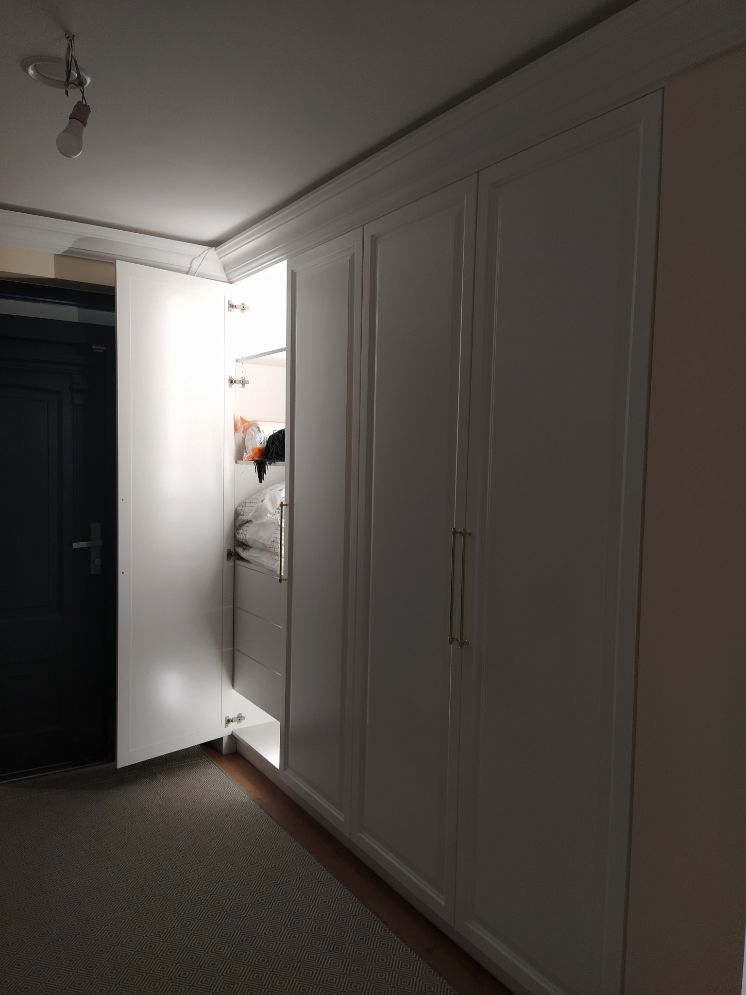 Biała szafa wnękowa z otwartymi drzwiami, oświetleniem wewnętrznym i złotymi uchwytami, widoczna zawartość szafy, w tle ciemne drzwi i wzorzysty dywan.