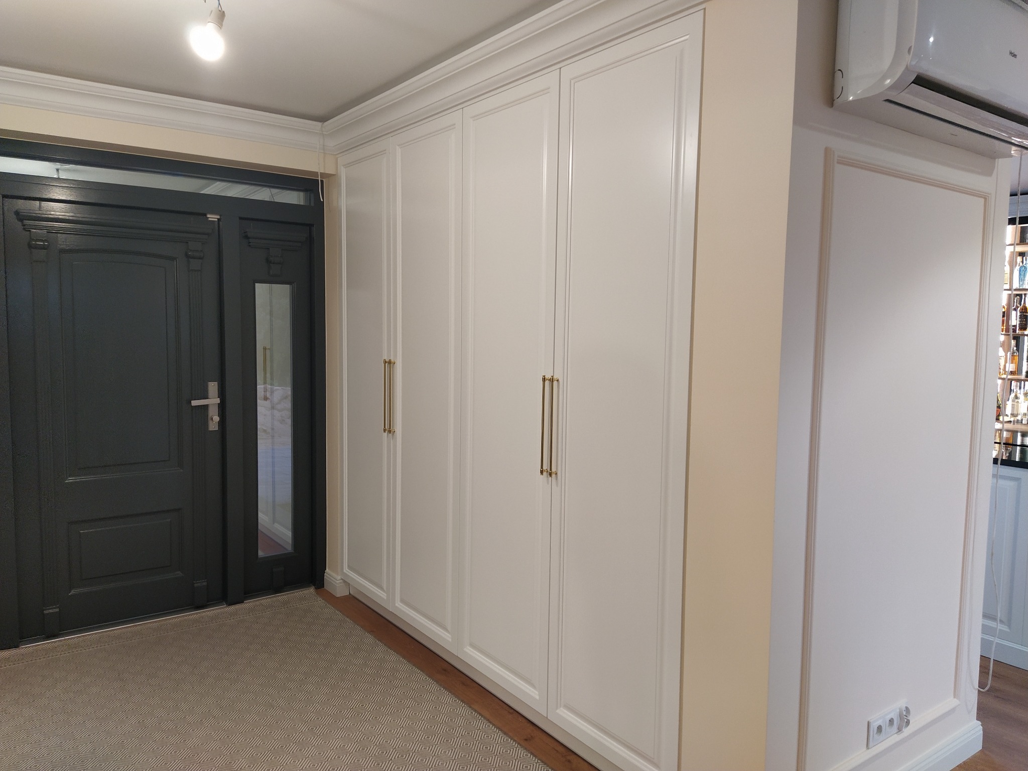 Biała szafa wnękowa z czterema drzwiami i złotymi uchwytami, dopasowana do wystroju wnętrza. Widoczna podłoga i ściana z gniazdkami.