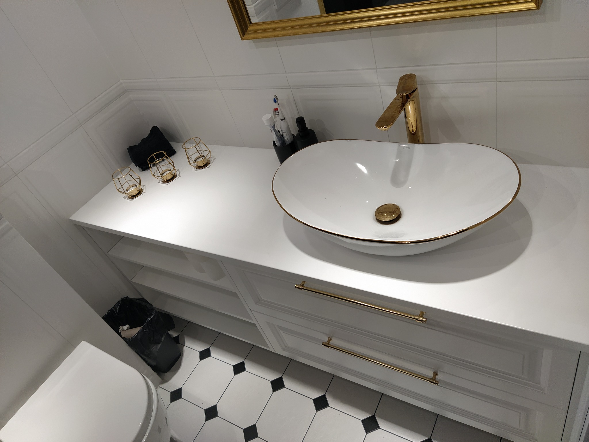Biała szafka łazienkowa z owalną umywalką i złotą armaturą, widok z góry, czarno-biała podłoga w tle, dekoracyjne świeczniki i złota rama lustra.