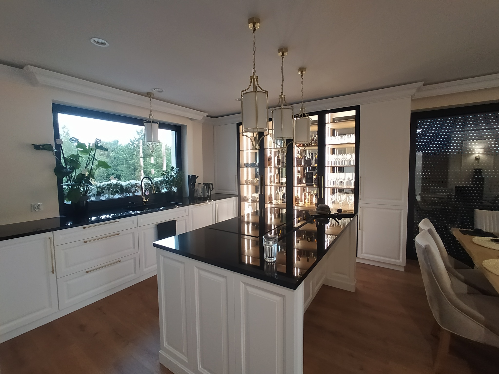 Kuchnia z wyspą i czarnym blatem, białe fronty szafek z frezami i złotymi uchwytami. Nad wyspą trzy złote lampy. W tle przeszklona szafa z alkoholem.