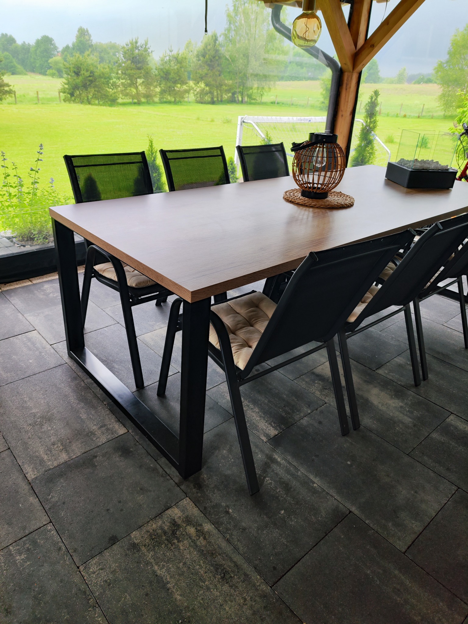 Stół z drewnianym blatem i metalowymi nogami w zadaszonej przestrzeni, z widokiem na zielony krajobraz. Nowoczesny design, naturalne światło.