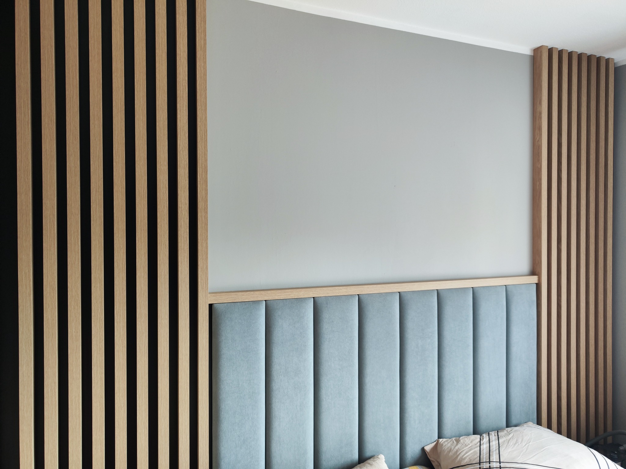 Nowoczesna aranżacja sypialni z wezgłowiem w pastelowym odcieniu, obudowanym drewnianymi lamelami. Minimalistyczny design i stonowana kolorystyka.