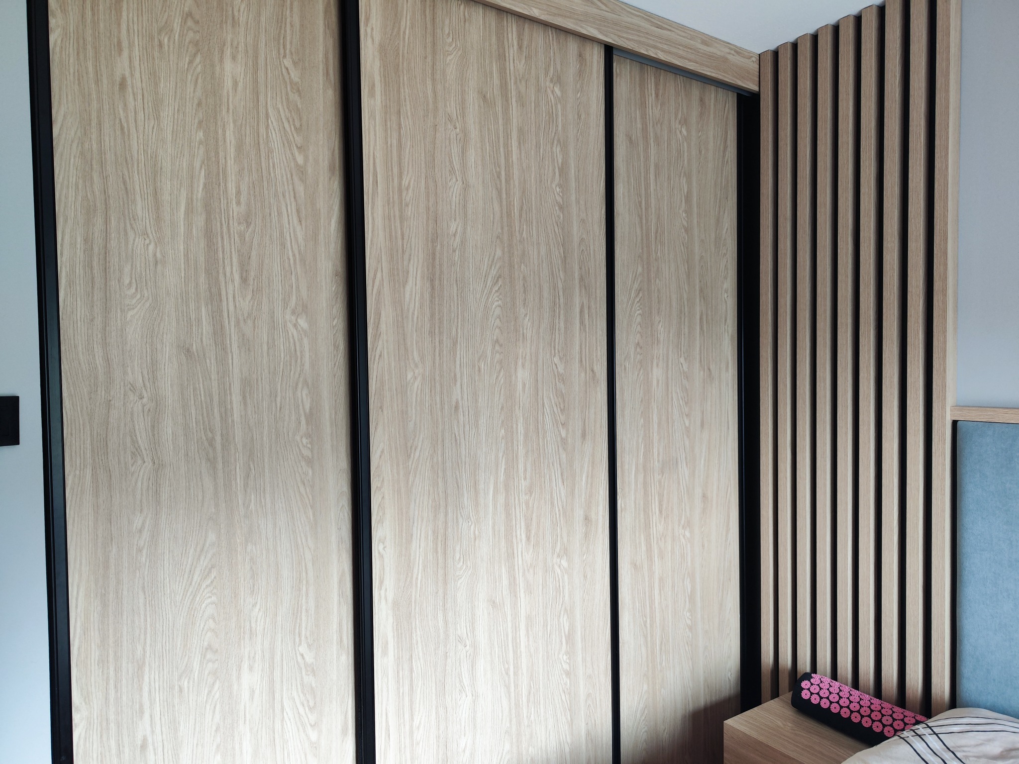 Nowoczesna szafa przesuwna z drewnopodobnymi frontami i czarnymi ramami, dopasowana kolorystycznie do lameli dekoracyjnych na ścianie w sypialni.