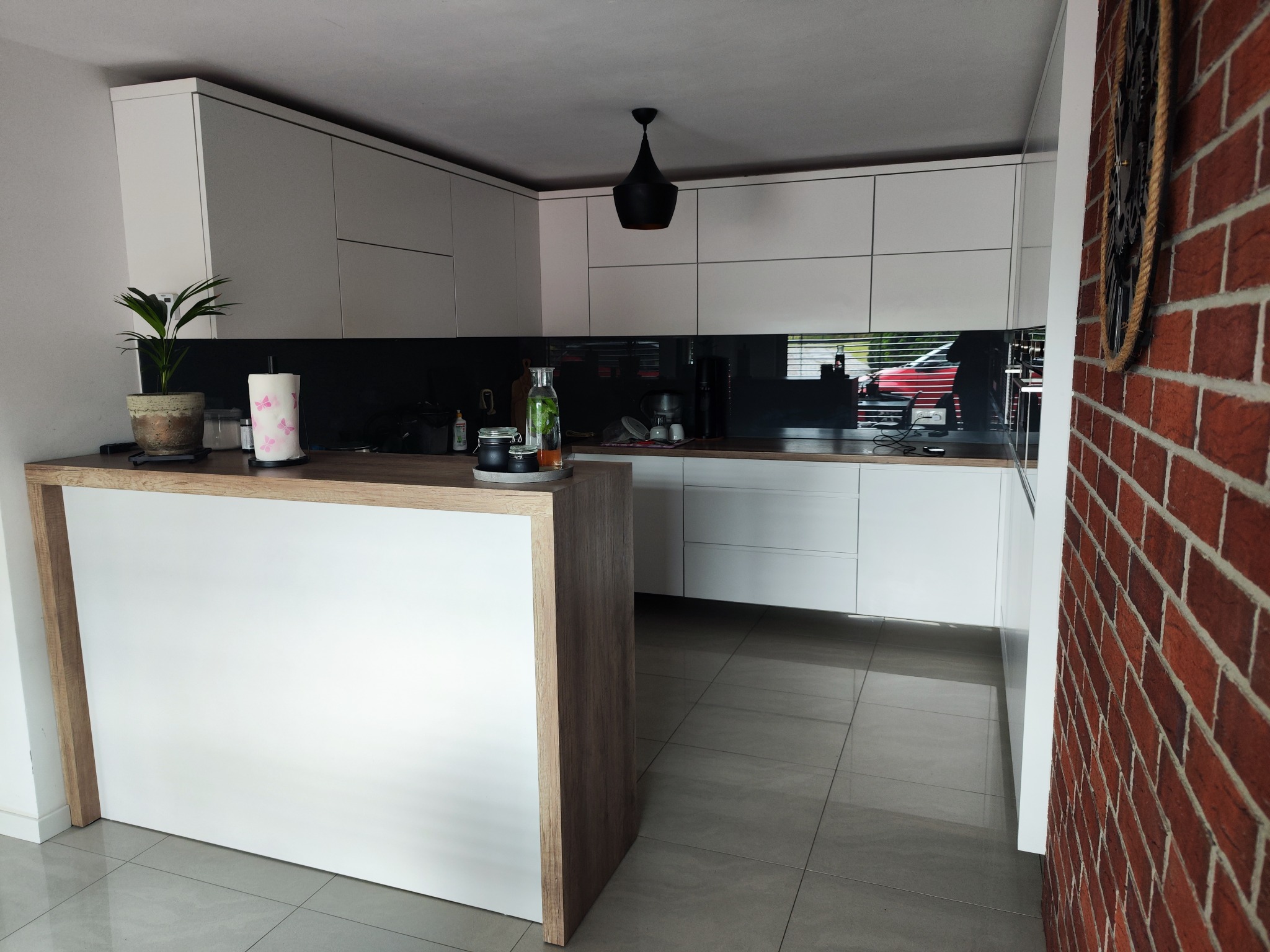 Nowoczesna kuchnia z białymi frontami i czarnym lacobelem, wyspa z blatem drewnopodobnym, ceglana ściana z dekoracją. Minimalistyczny design.