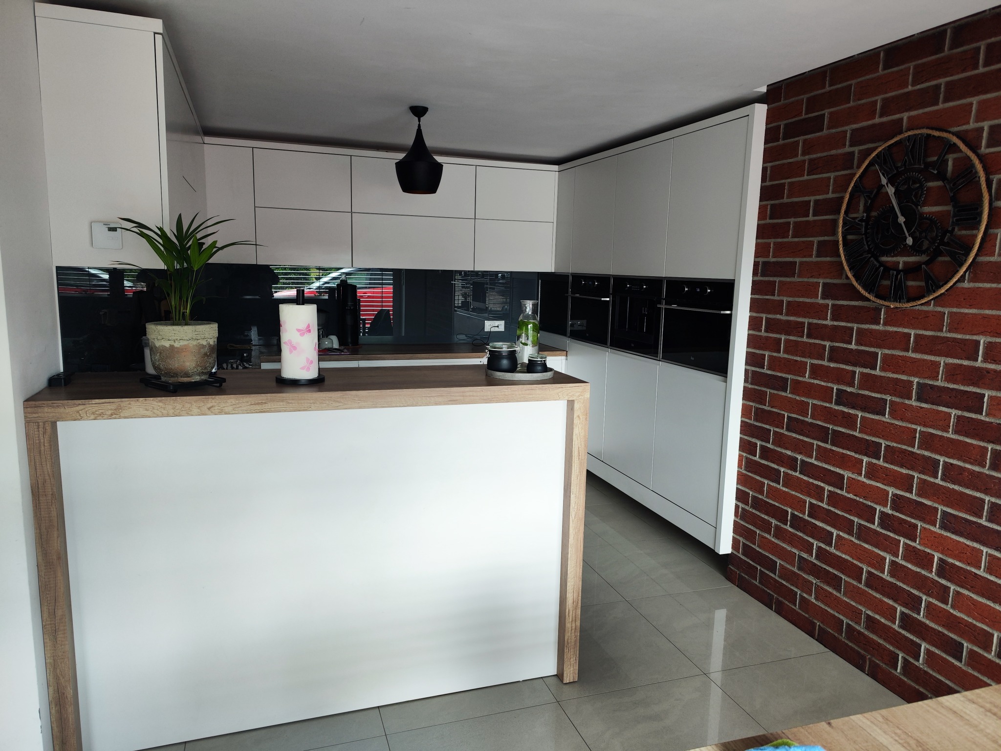Nowoczesna kuchnia z białymi szafkami, czarnym blatem i ceglaną ścianą z zegarem. Minimalistyczny design, oświetlenie punktowe i roślina doniczkowa.