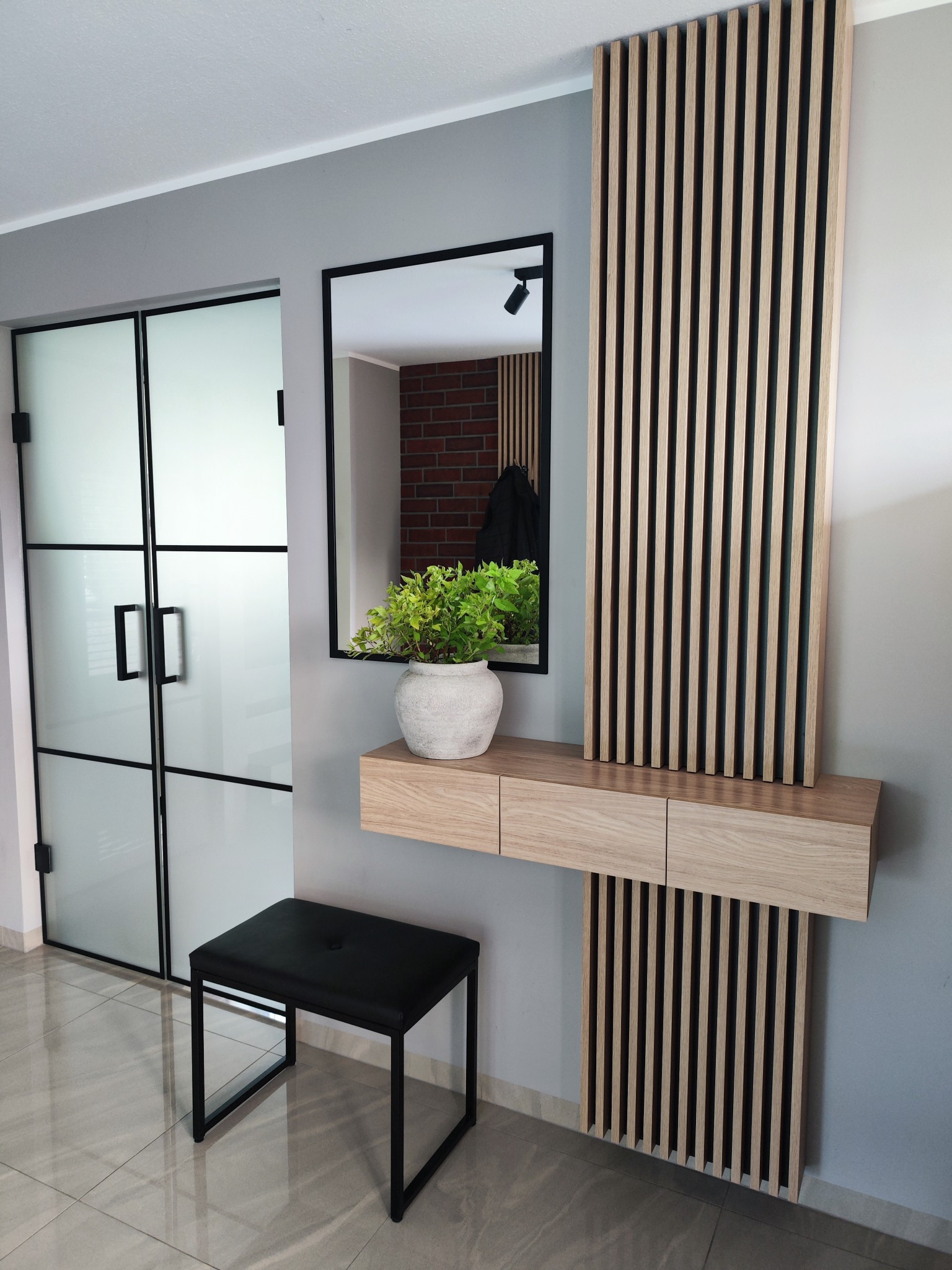 Nowoczesna szafka z lustrem i dekoracyjnymi lamelami w przedpokoju. Czarny taboret, drzwi z matowego szkła z czarnymi szprosami, płytki podłogowe.