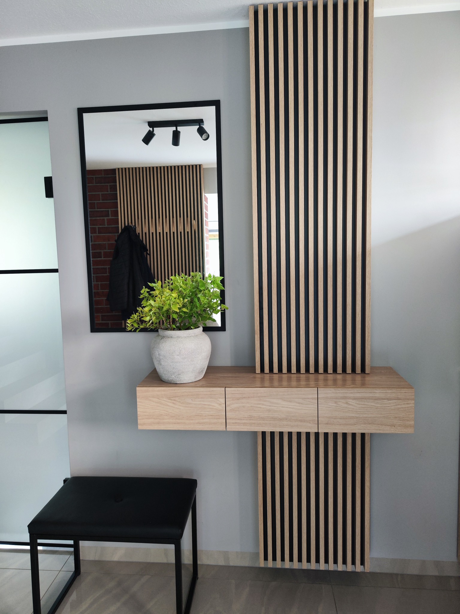 Nowoczesna konsola z lustrem i lamelami ściennymi w przedpokoju. Jasne drewno, czarne akcenty, minimalistyczny design. Roślina w doniczce dodaje naturalnego akcentu.