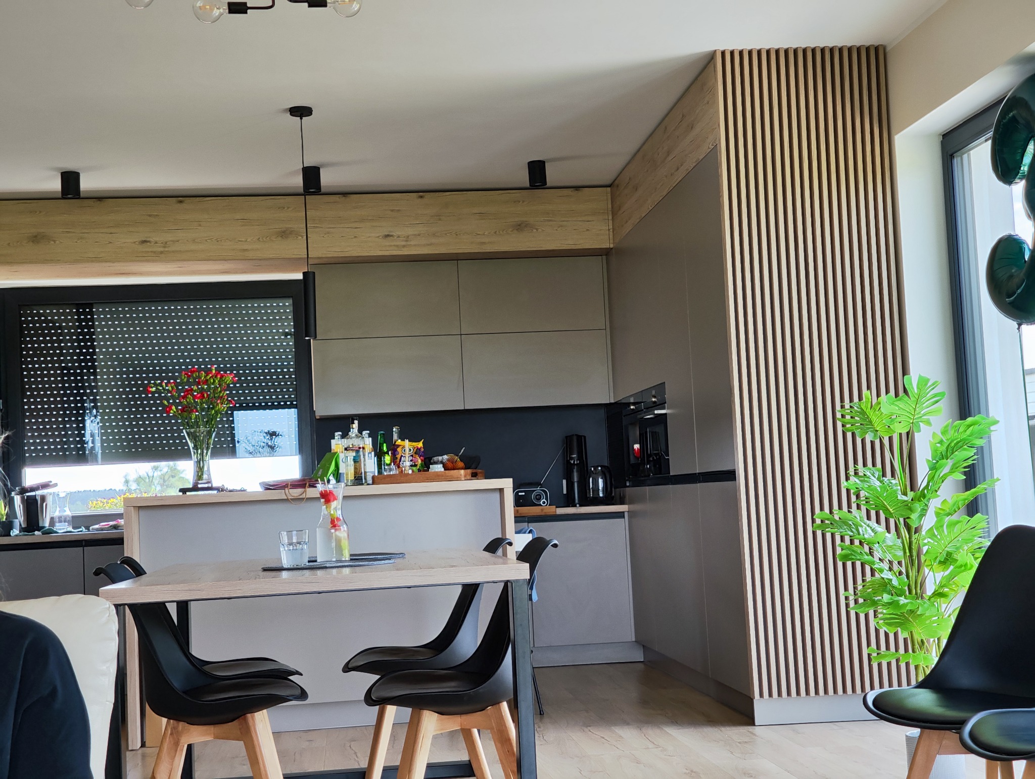 Nowoczesna kuchnia z drewnianymi lamelami na ścianie, stół z czarnymi krzesłami, kwiaty w wazonie, minimalistyczne oświetlenie i zabudowa kuchenna w odcieniach szarości.