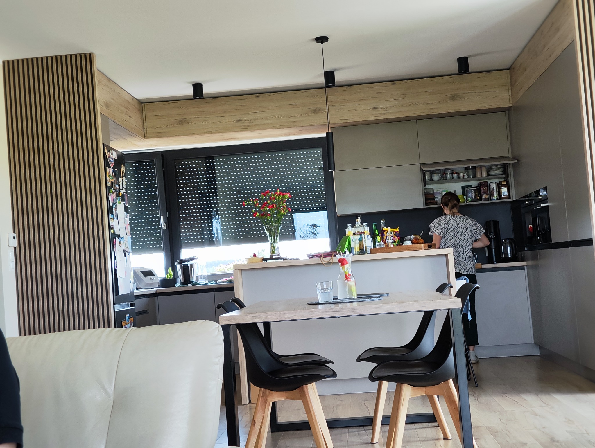 Nowoczesna kuchnia z wyspą i drewnianymi akcentami, widok na stół z krzesłami, osoba w tle przygotowująca posiłek, minimalistyczny design.