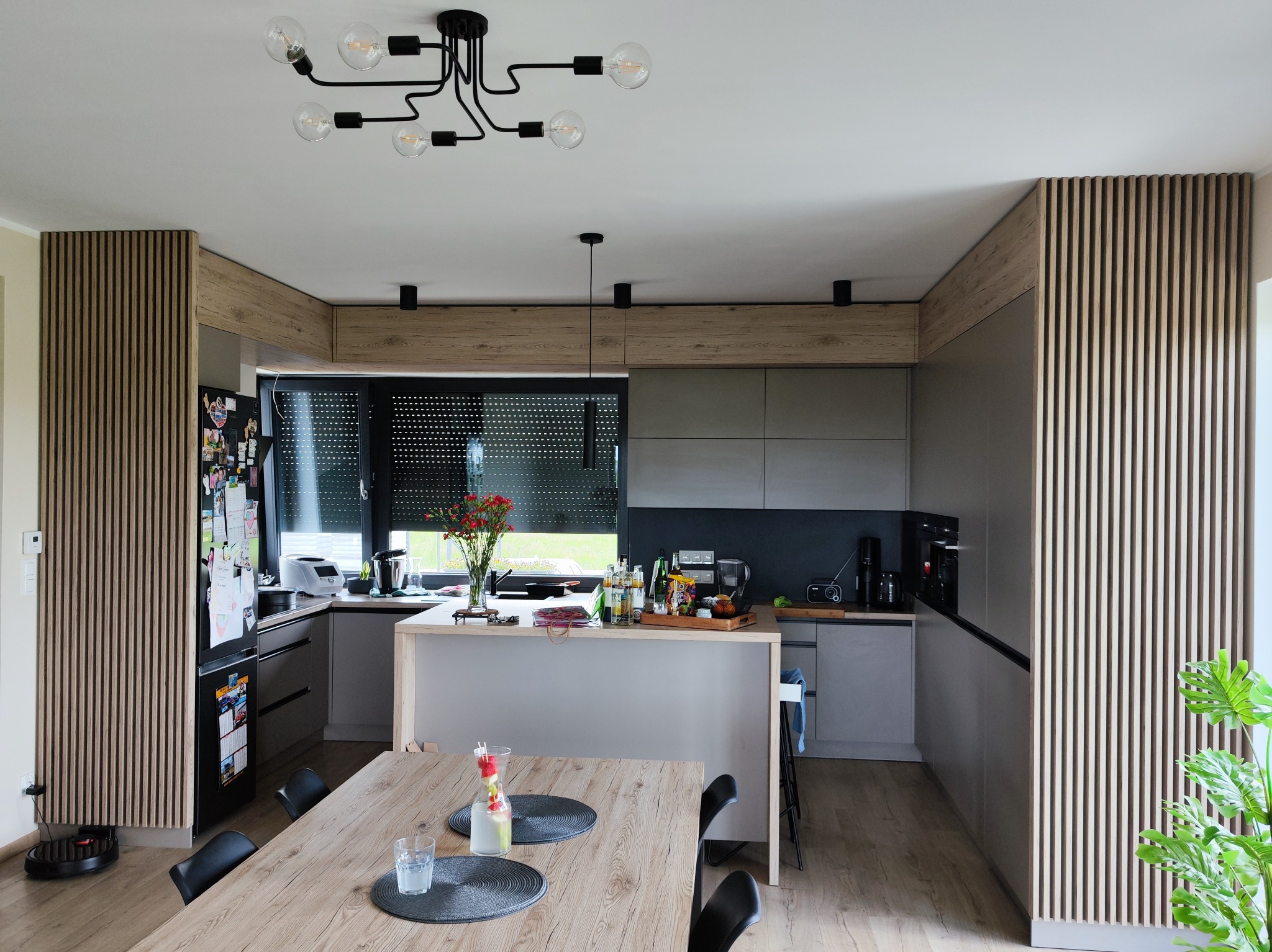 Nowoczesna kuchnia z szarymi szafkami, drewnianymi panelami ściennymi i wyspą. Na stole napój w dzbanku. Czarny żyrandol z żarówkami Edison.