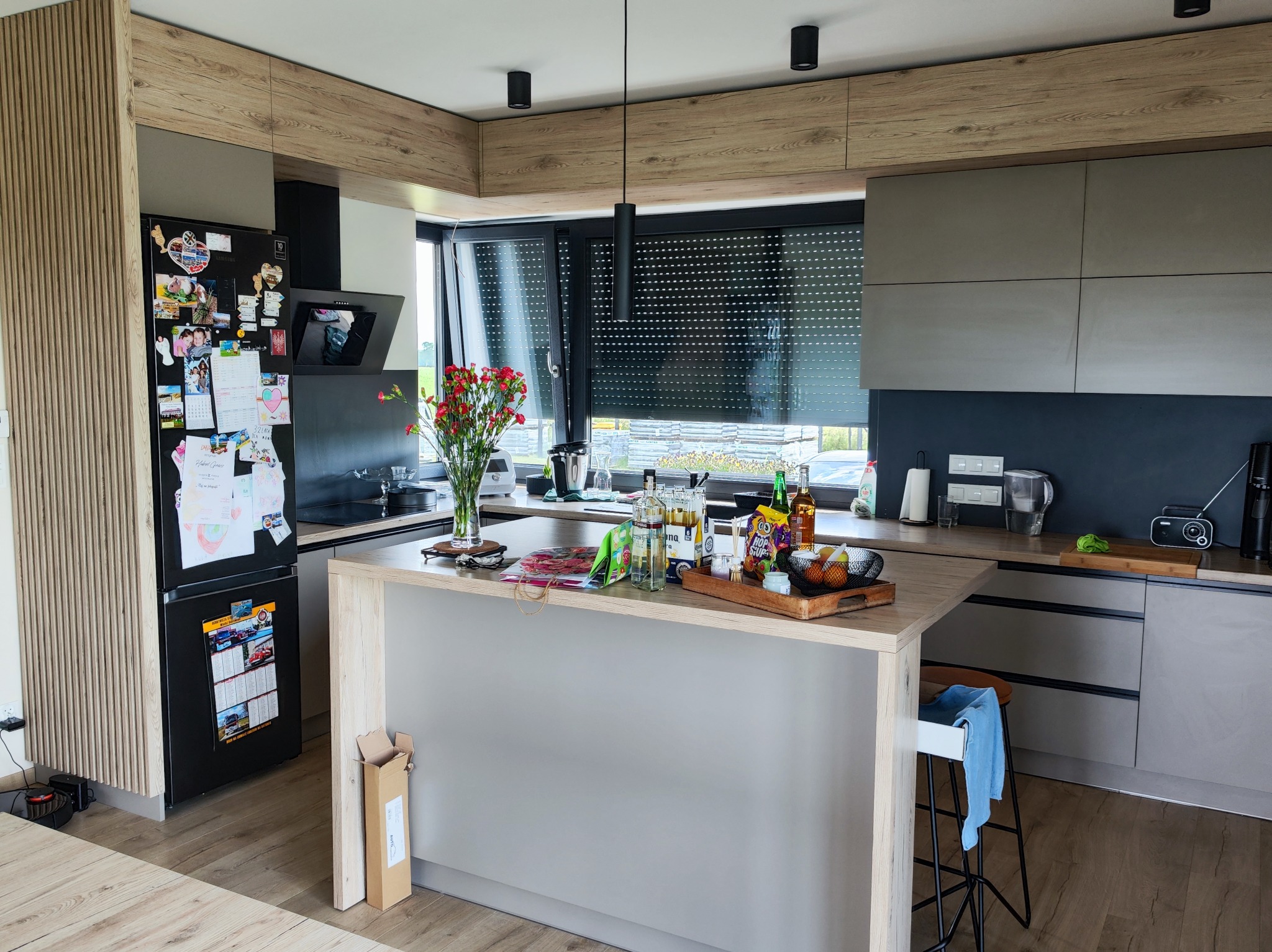 Nowoczesna kuchnia z wyspą, szarymi frontami i drewnianymi akcentami. Na wyspie kwiaty i akcesoria. Na lodówce magnesy i kartki. Widok na zewnątrz przez okno.