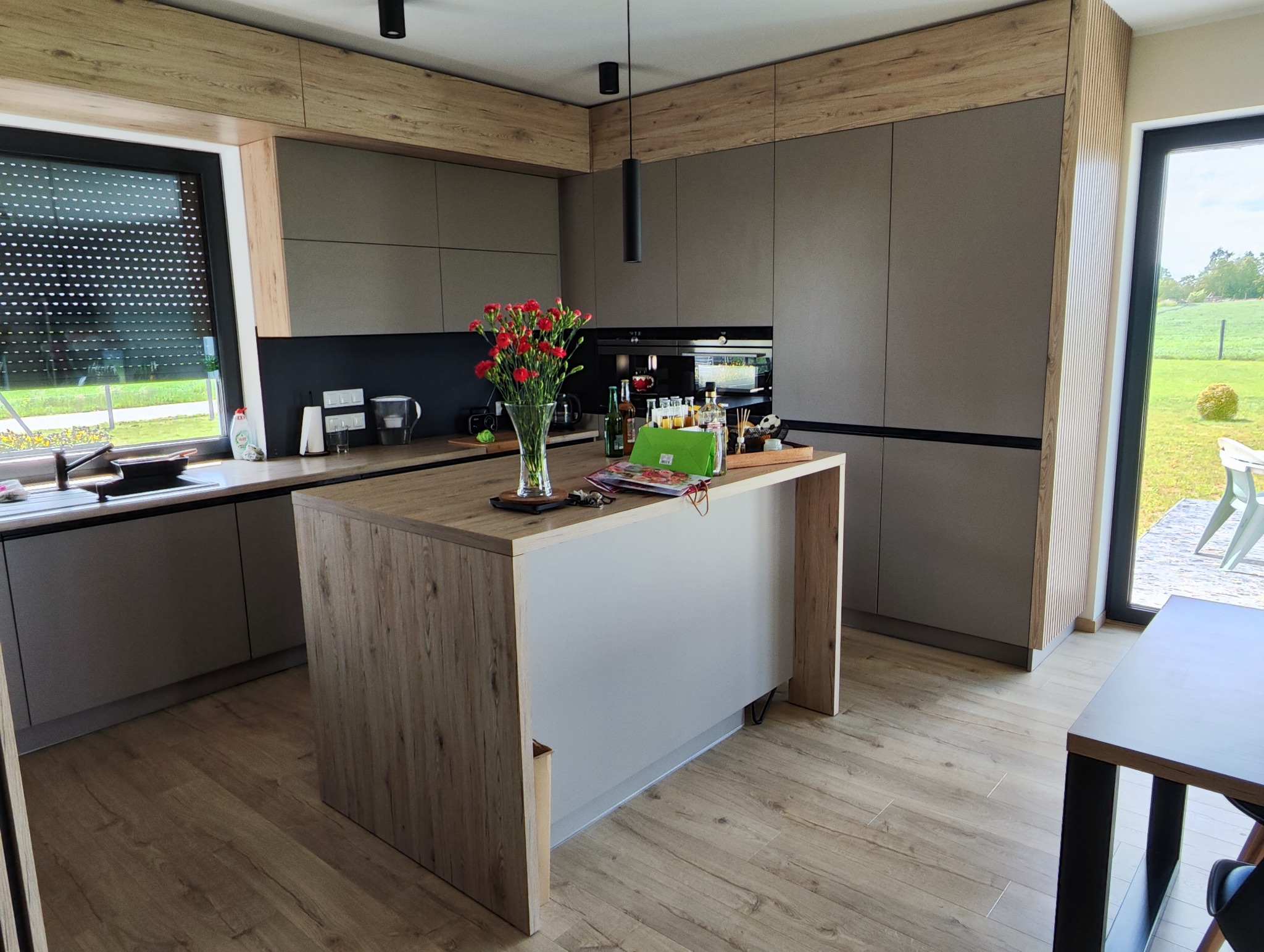 Nowoczesna kuchnia z wyspą i drewnianymi akcentami. Szare fronty szafek i blaty imitujące drewno tworzą spójną całość. Widok na zieleń za oknem.